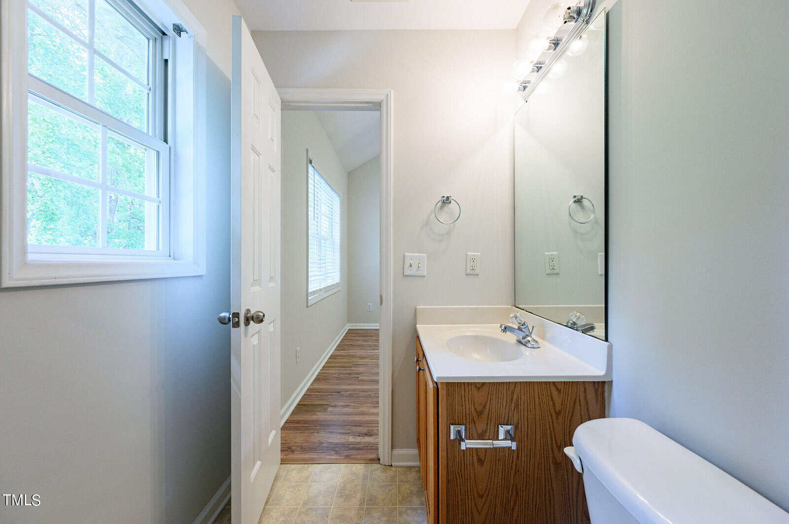 A small bathroom with a wooden vanity, a white countertop, and a sink. There's a mirror with lights above it, a window on the left, and a door leading to a room with wooden flooring. A toilet is visible in the foreground.