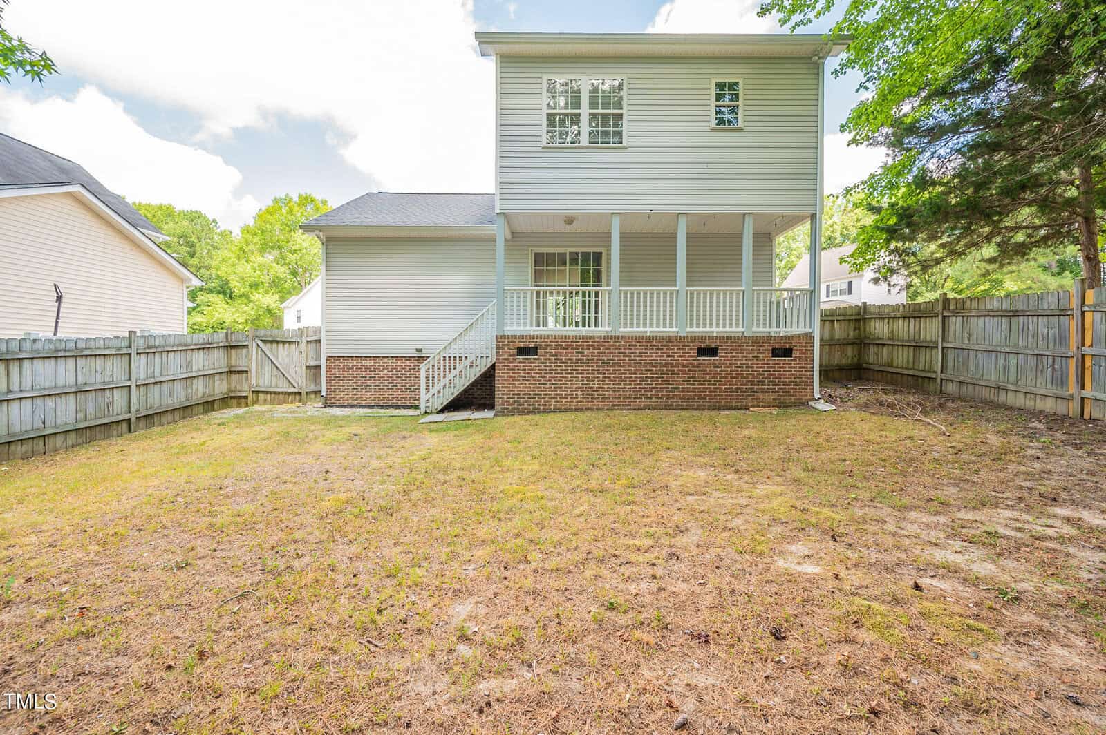 A two-story house with light blue siding and white trim, featuring a raised porch with stairs leading down to a fenced backyard. The yard is partially covered in grass, surrounded by wooden fencing and trees.