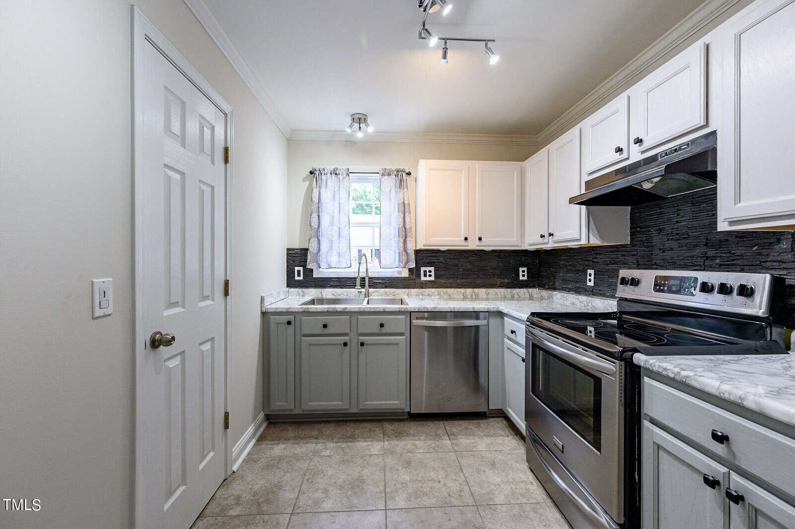 A modern kitchen featuring white upper cabinets, gray lower cabinets, and stainless steel appliances, including a stove and dishwasher. A window above the sink lets in natural light, and a track light fixture illuminates the space.