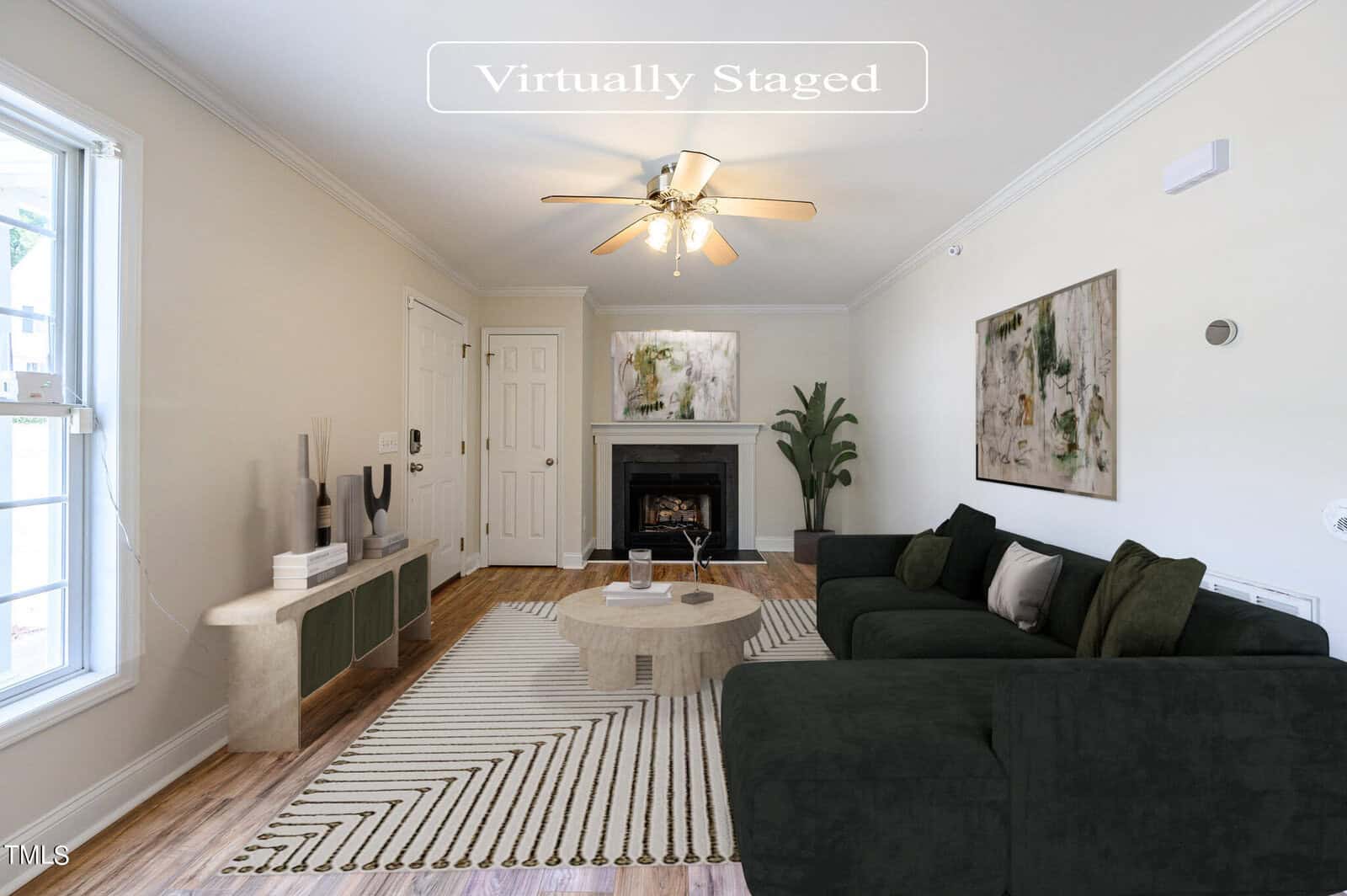 A virtually staged living room featuring a green sectional sofa, round wooden table, abstract rug, and a fireplace. A tall plant and modern decor items are placed along the walls. A ceiling fan is centered above. Natural light enters through the window.