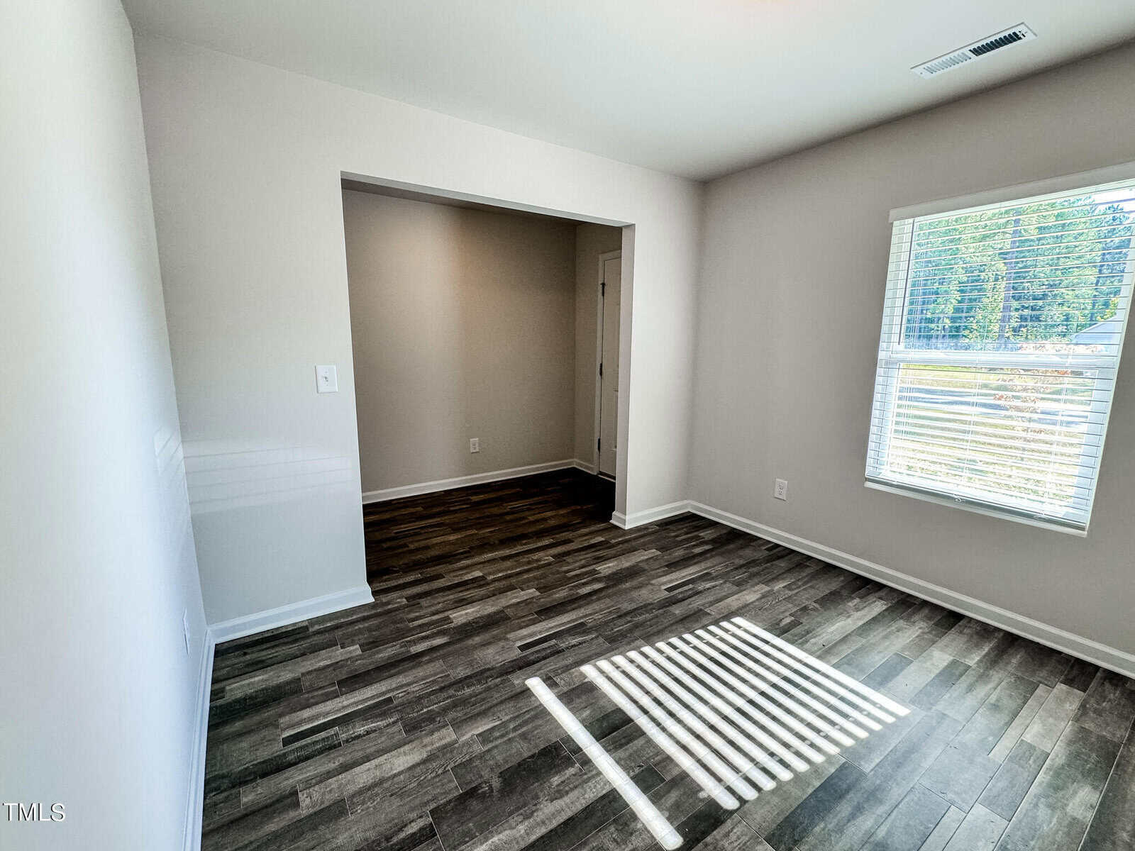 A room with dark wood laminate flooring, light beige walls, and a window allowing natural light to stream in. Adjacent to it is a recessed area or small room visible through an open doorway, leading to another door on the far wall.