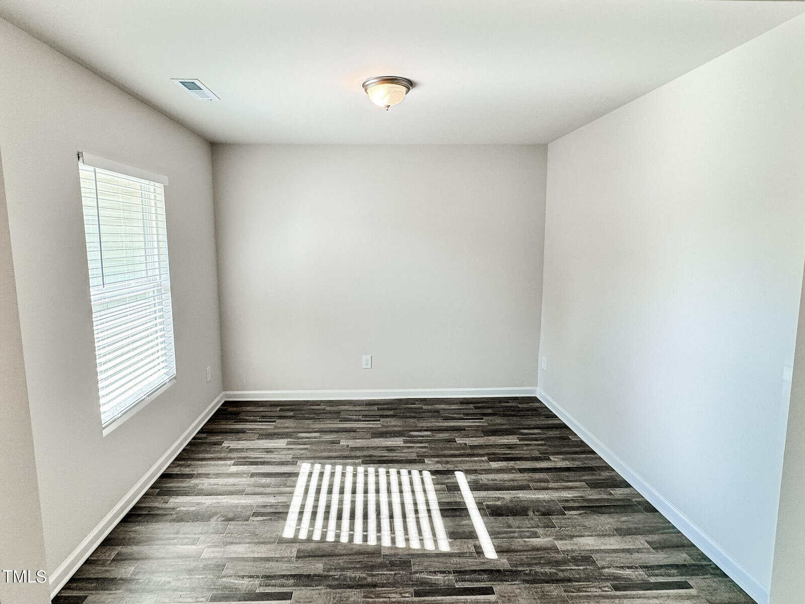 A small, empty room with light gray walls and a dark wood floor. A single ceiling light fixture is centered on the ceiling. A large window with blinds allows sunlight to stream in, casting shadows on the floor. The room has a bright and clean appearance.