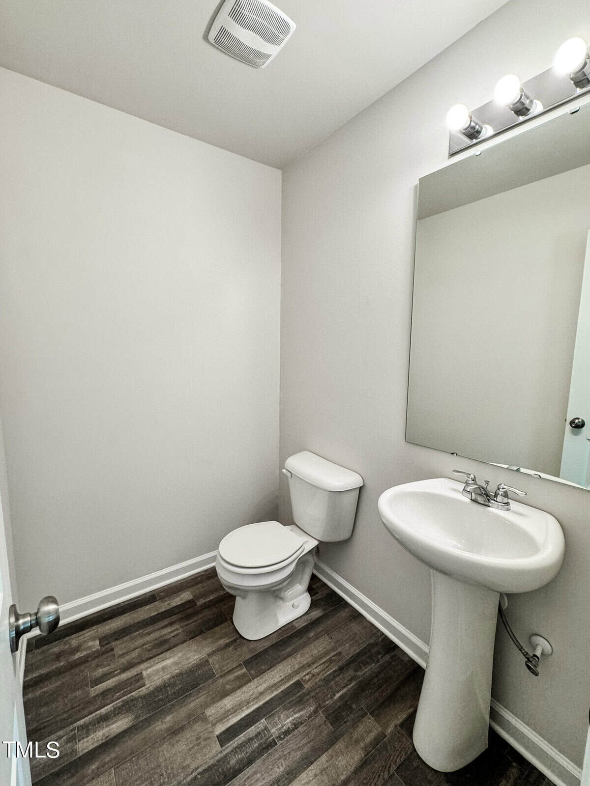 A small bathroom with a white toilet and a white pedestal sink. Above the sink, there is a large mirror with three light fixtures. The floor is covered in dark wood-like tiles. The walls are plain white, with no additional decorations or accessories.