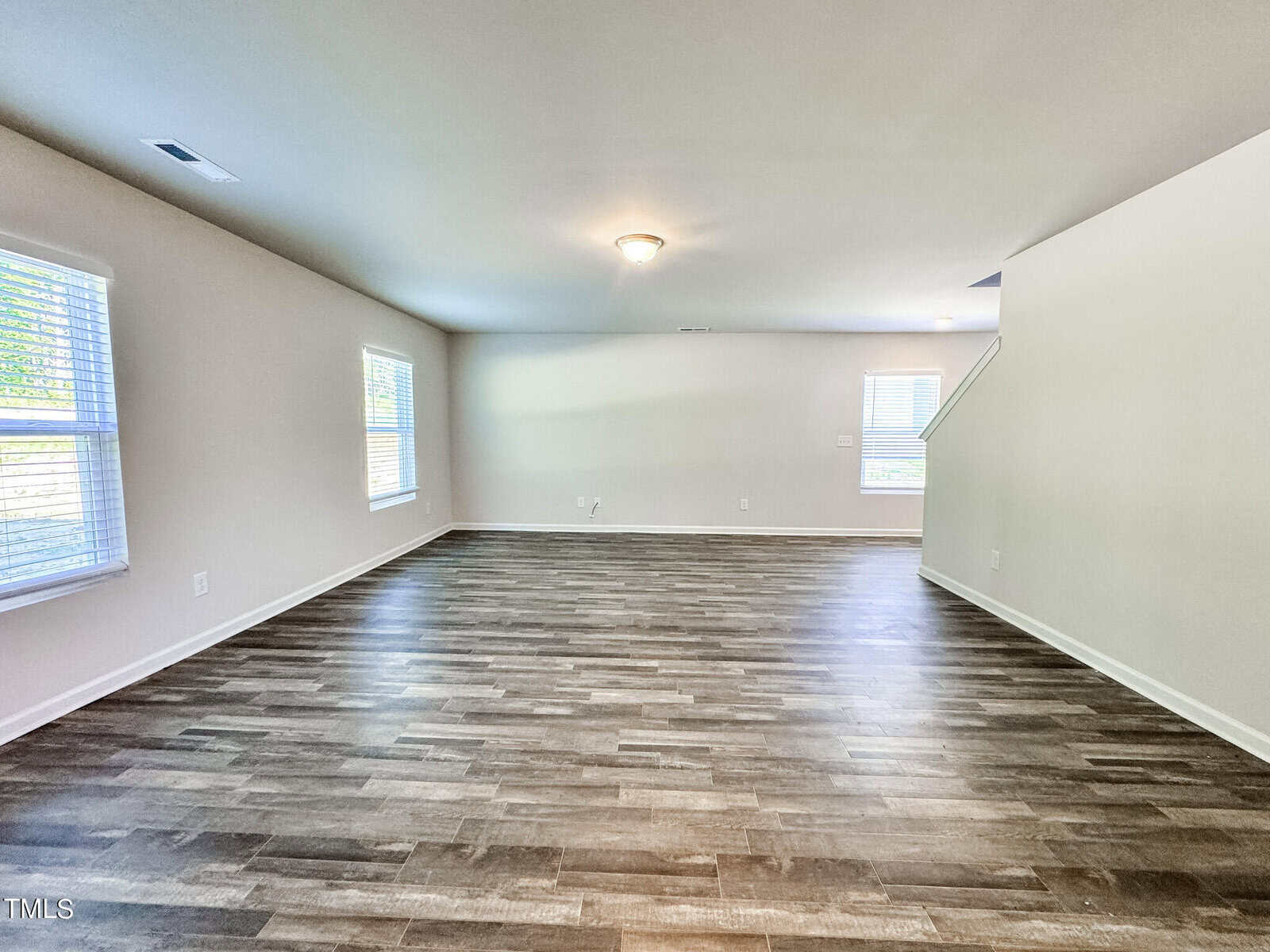 A spacious empty living room with light brown wood flooring, beige walls, and recessed lighting. Large windows on two walls allow natural light to fill the space. Stairs in the background lead to the upper level.