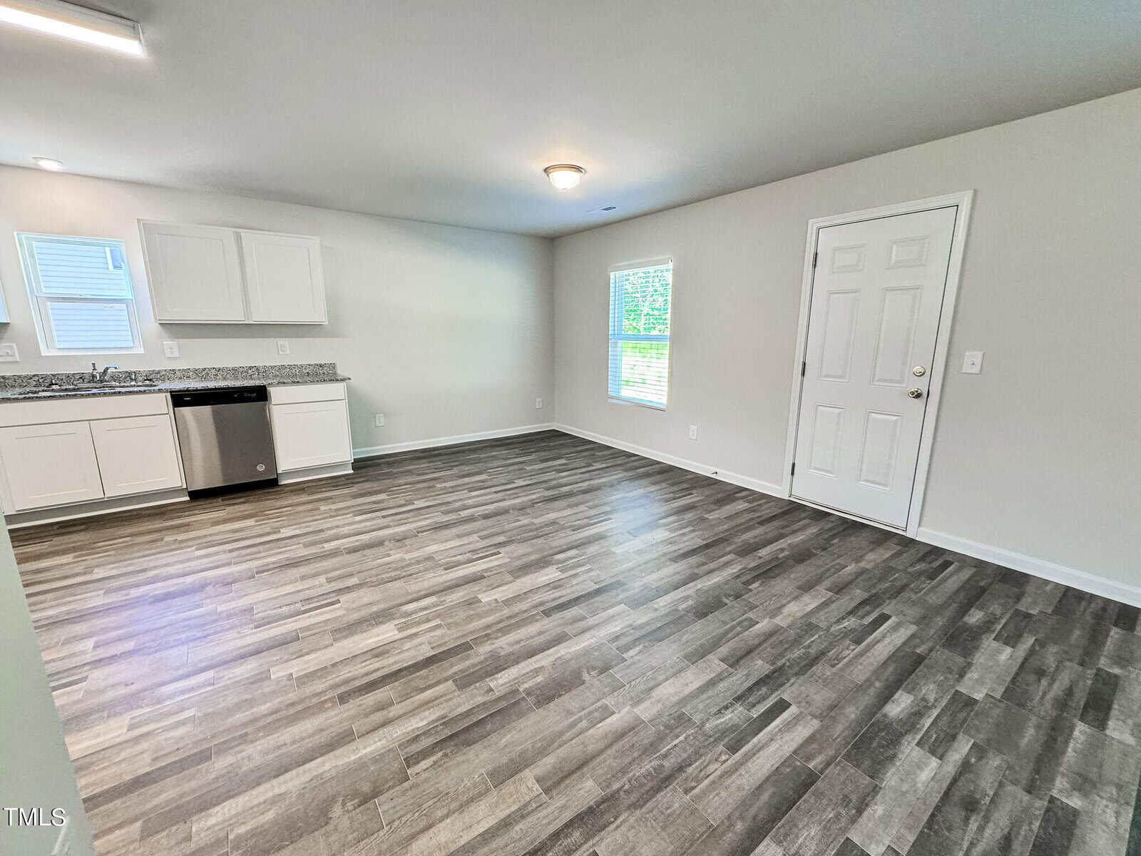 A photo of an empty room with light-colored walls and wood-patterned flooring. The room features a kitchenette with white cabinets, a stainless steel dishwasher, and a view window. A white door is present on the right side, along with a window letting in natural light.