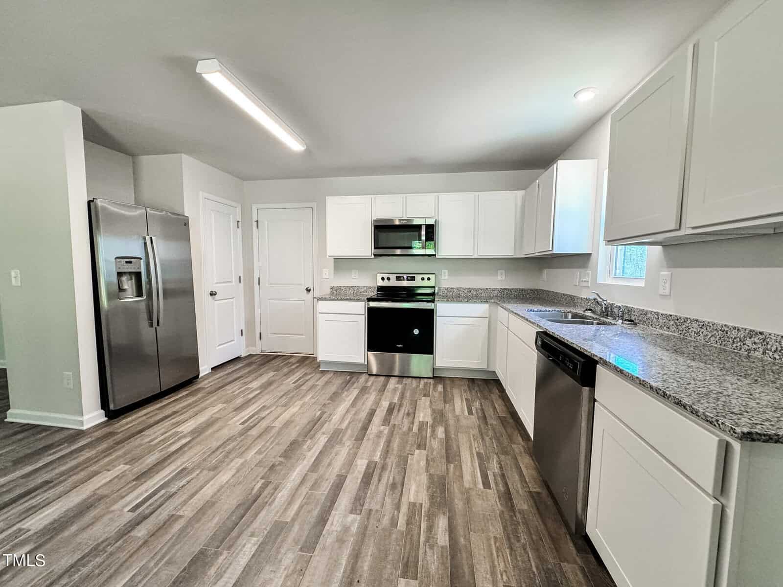 A modern kitchen with white cabinets, stainless steel appliances including a refrigerator, oven, microwave, and dishwasher. Granite countertops and a wood-patterned floor are also featured. A window above the sink provides natural light.