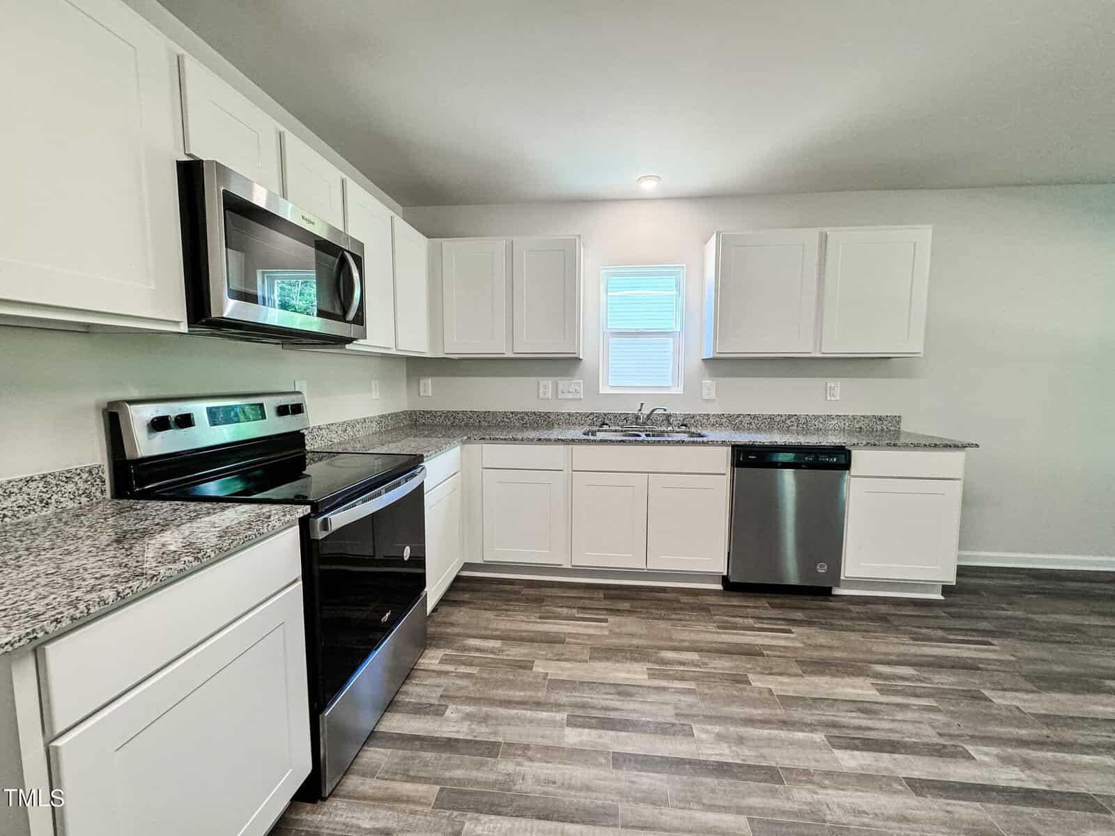 A modern kitchen features white cabinets and a speckled granite countertop. Stainless steel appliances, including a microwave, electric stove, and dishwasher, are installed. The room has wood-look vinyl flooring and a small window above the sink.