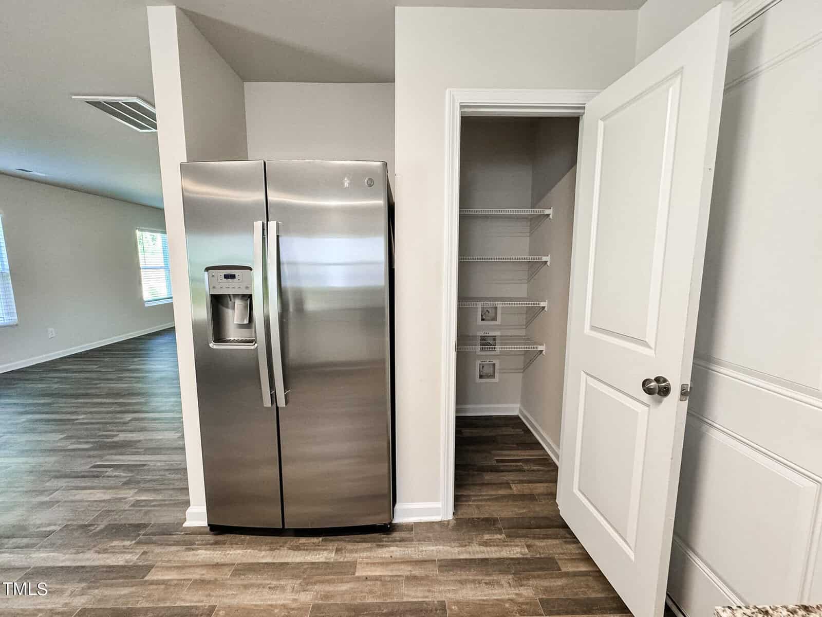 A modern kitchen features a stainless steel side-by-side refrigerator with a water and ice dispenser. Adjacent is an open pantry with built-in shelves. The room has white walls and wood-look vinyl flooring, leading into a bright living space.
