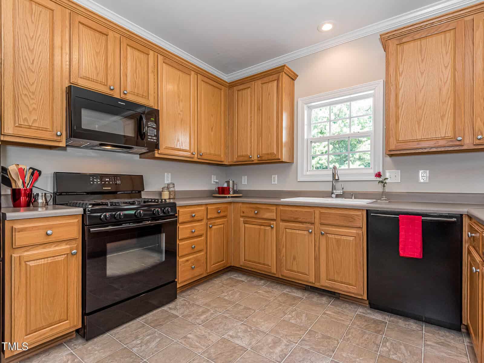 A modern kitchen with wooden cabinets and tile flooring. It features a black stove and microwave, black dishwasher, and a stainless steel sink. A small window above the sink lets in natural light. A red towel hangs on the dishwasher handle.