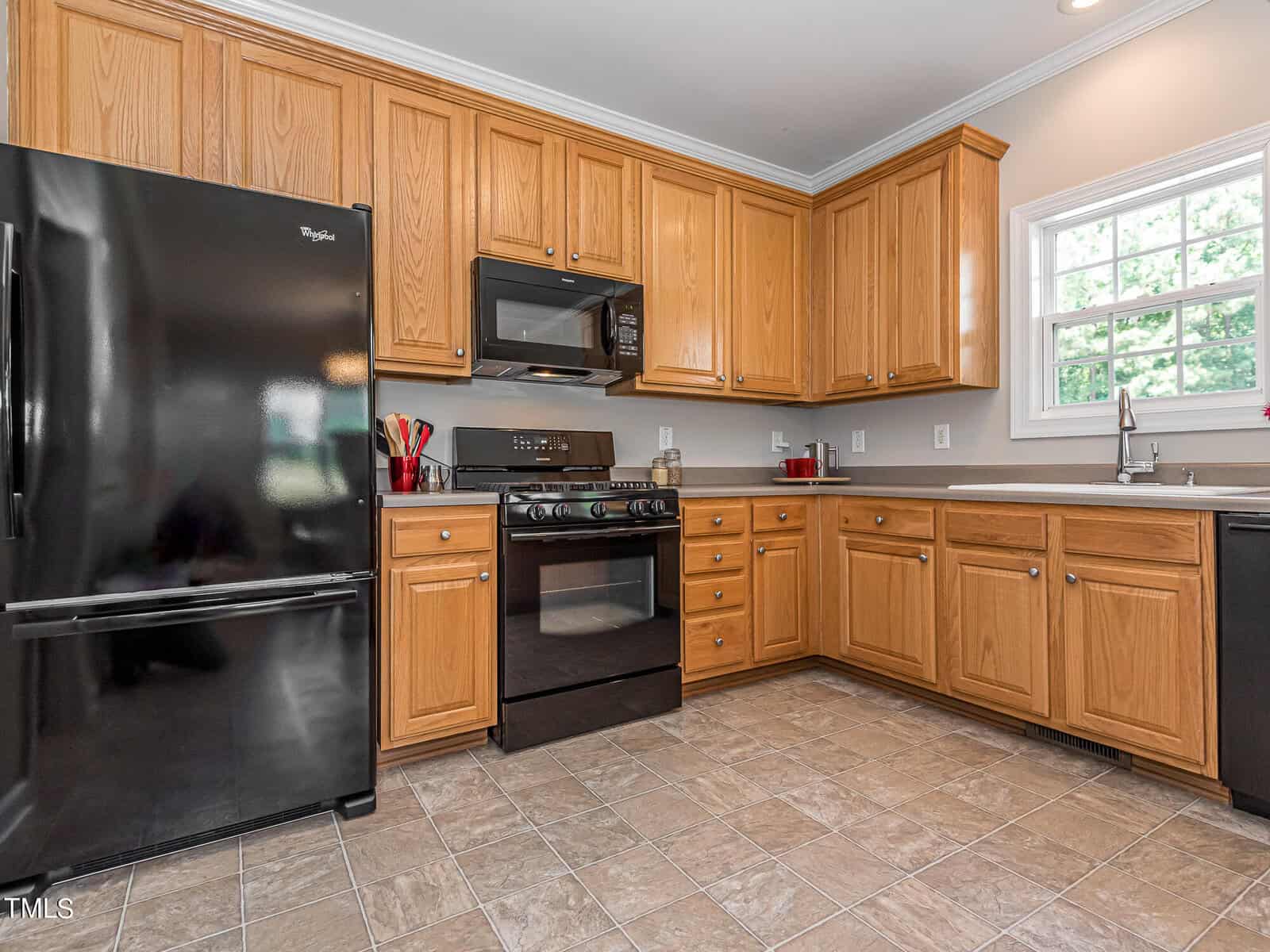 A modern kitchen featuring wooden cabinets, a black refrigerator, stove, and microwave. The countertop holds a few kitchen items. A window on the right allows natural light to brighten the space. The floor is tiled in a light beige color.
