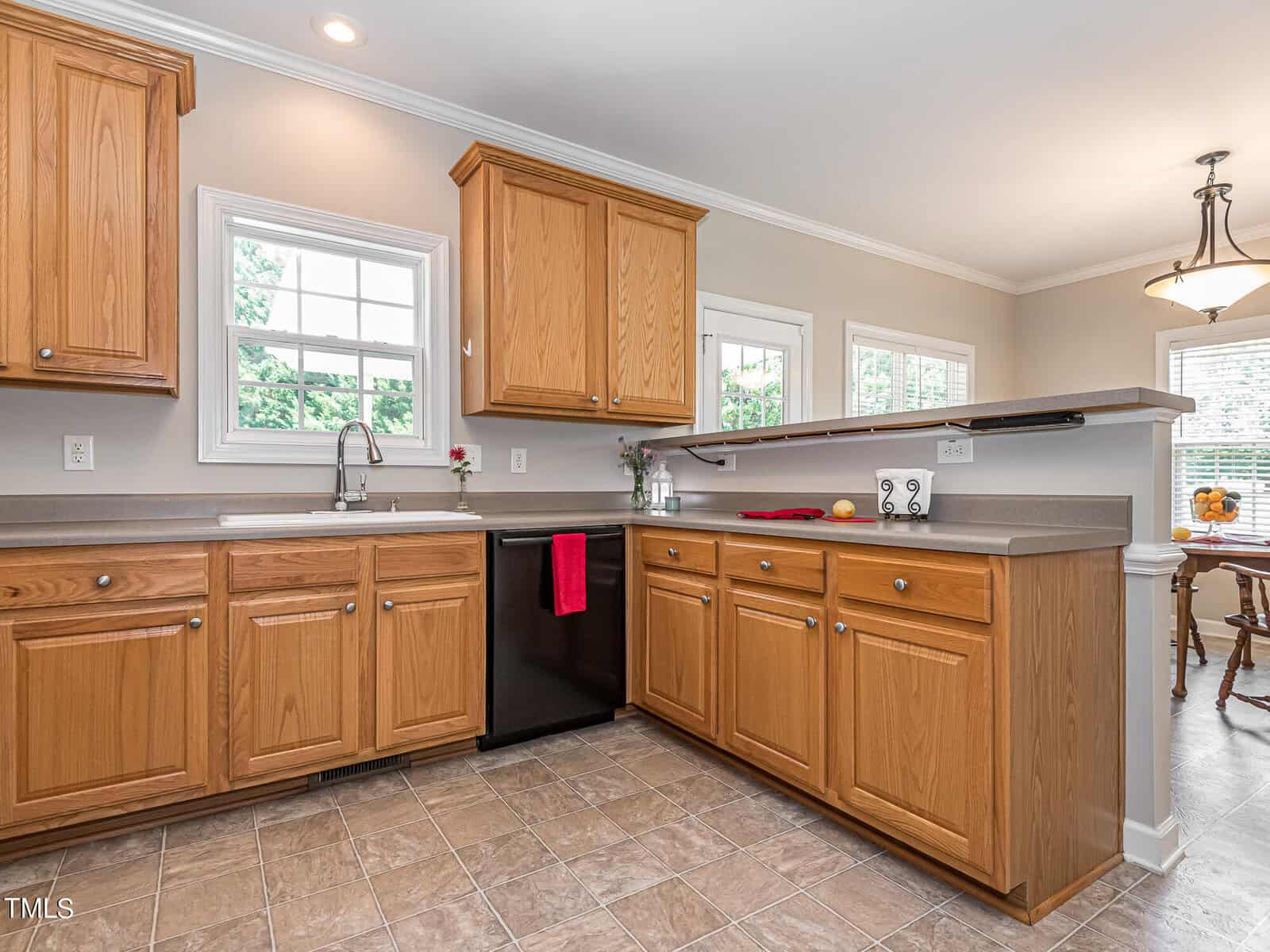 A spacious kitchen with wooden cabinets, a black dishwasher, and a dual sink under a window. The countertop holds a plant, a red towel, and a bowl of lemons. Adjacent is a dining area with a wooden table and chairs, lit by a hanging light fixture.