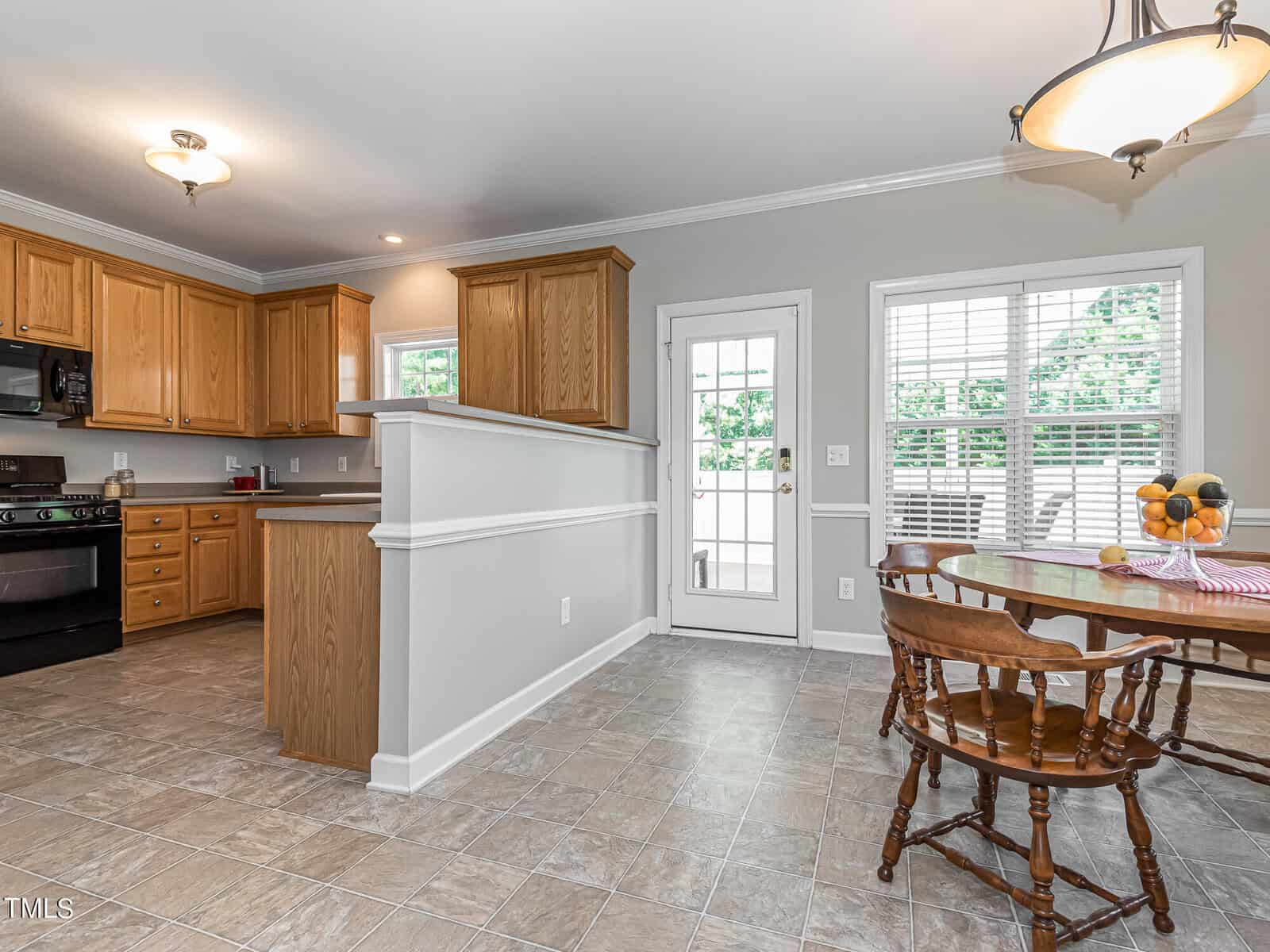 A bright kitchen with wooden cabinets, a black stove, and a small dining table. The room features light gray walls, a tiled floor, and a door with windows leading outside. The table is set with a bowl of fruit.