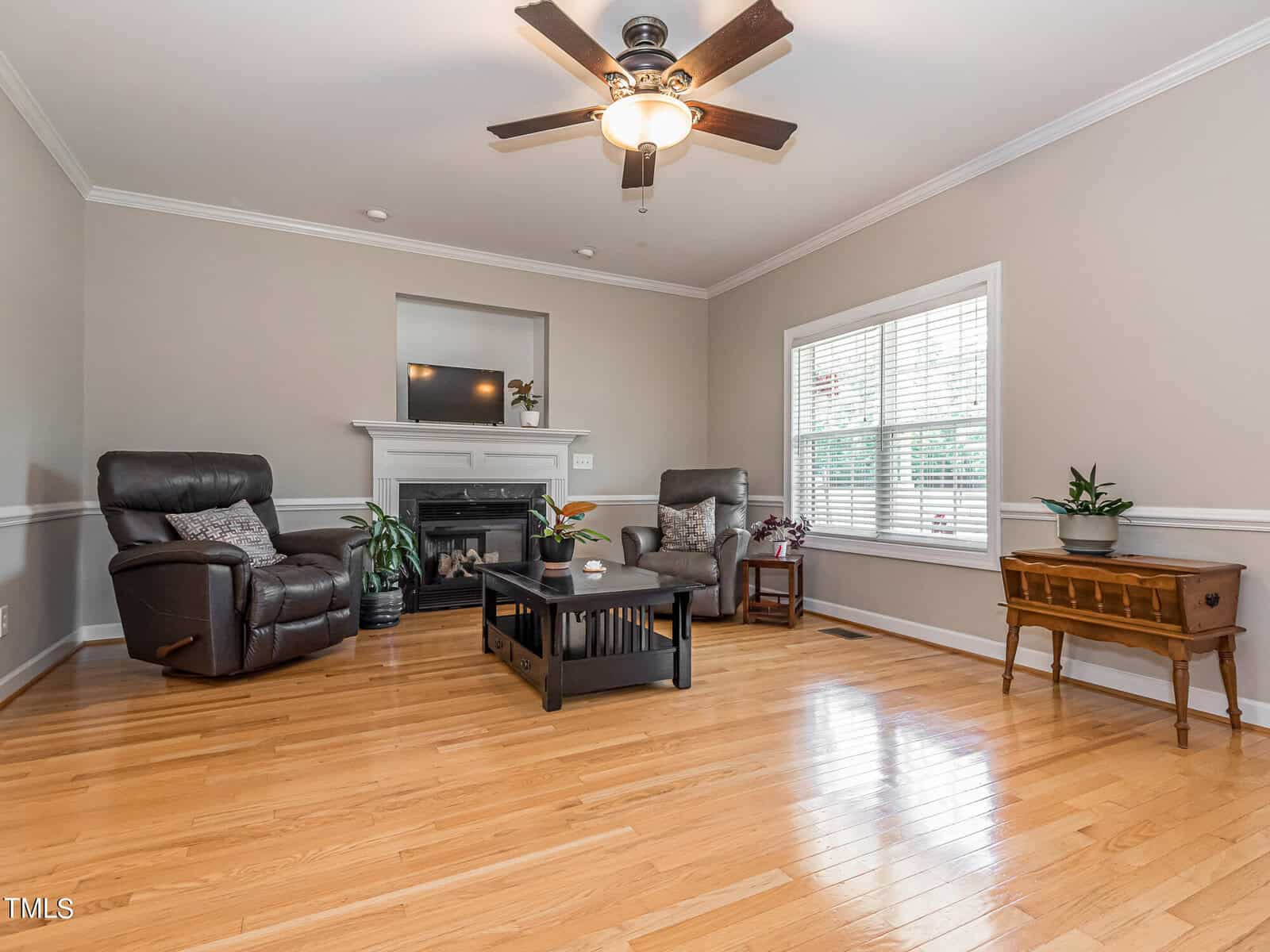 A spacious living room with light hardwood floors and neutral walls. Two recliners face a black fireplace with a white mantel. A dark coffee table is centered on a rug. A ceiling fan hangs from above, and a window lets in natural light.