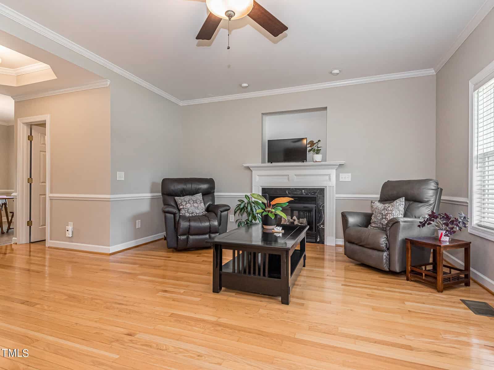 A cozy living room featuring two gray recliners, a black coffee table with plants, and a fireplace with a white mantel. The room has hardwood floors, neutral walls, and a ceiling fan. Natural light enters through a window on the right.