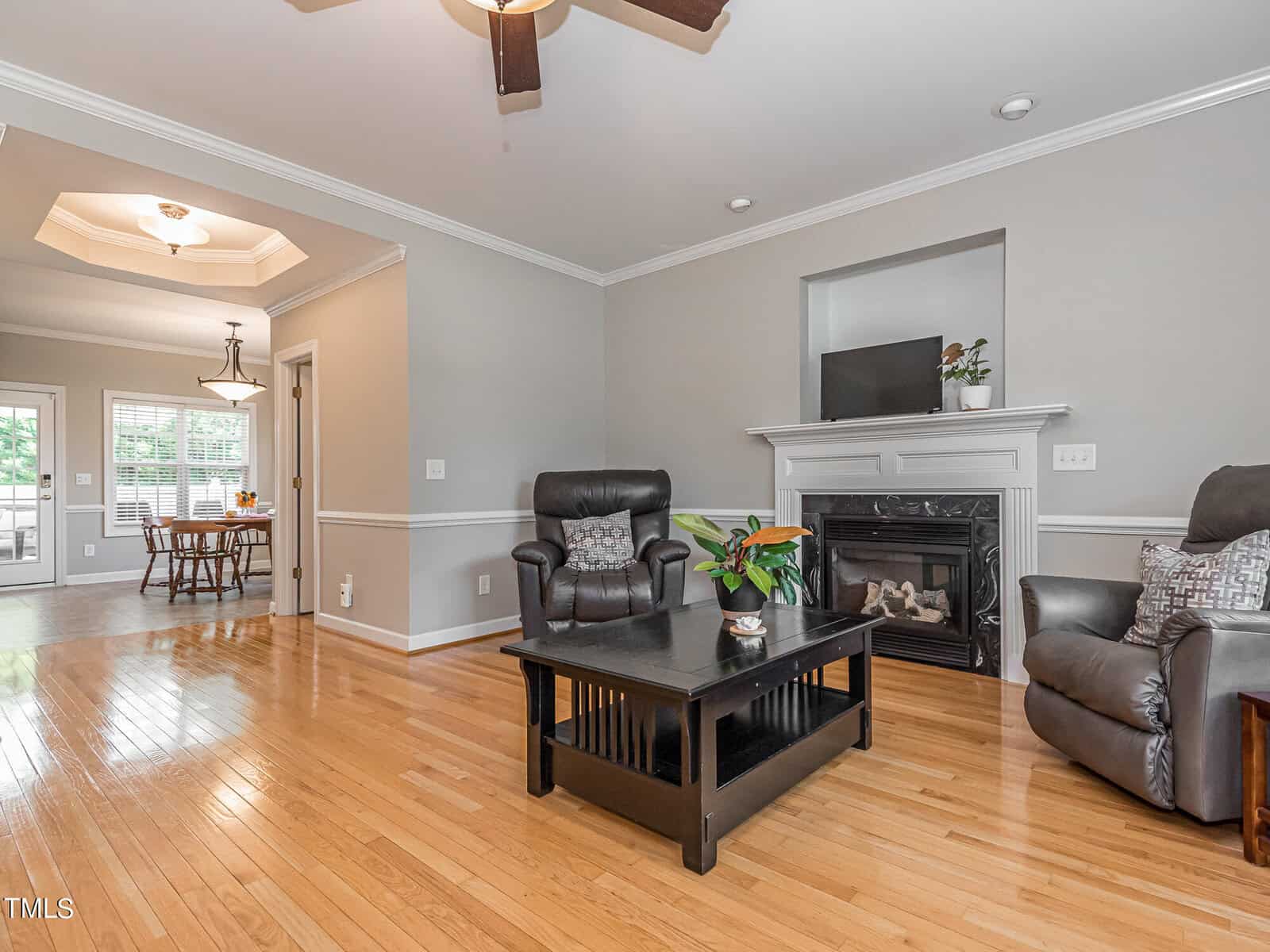Living room with wooden floors, beige walls, and a ceiling fan. Features a black coffee table with a plant, two dark leather chairs, and a fireplace. A dining area is visible in the background.