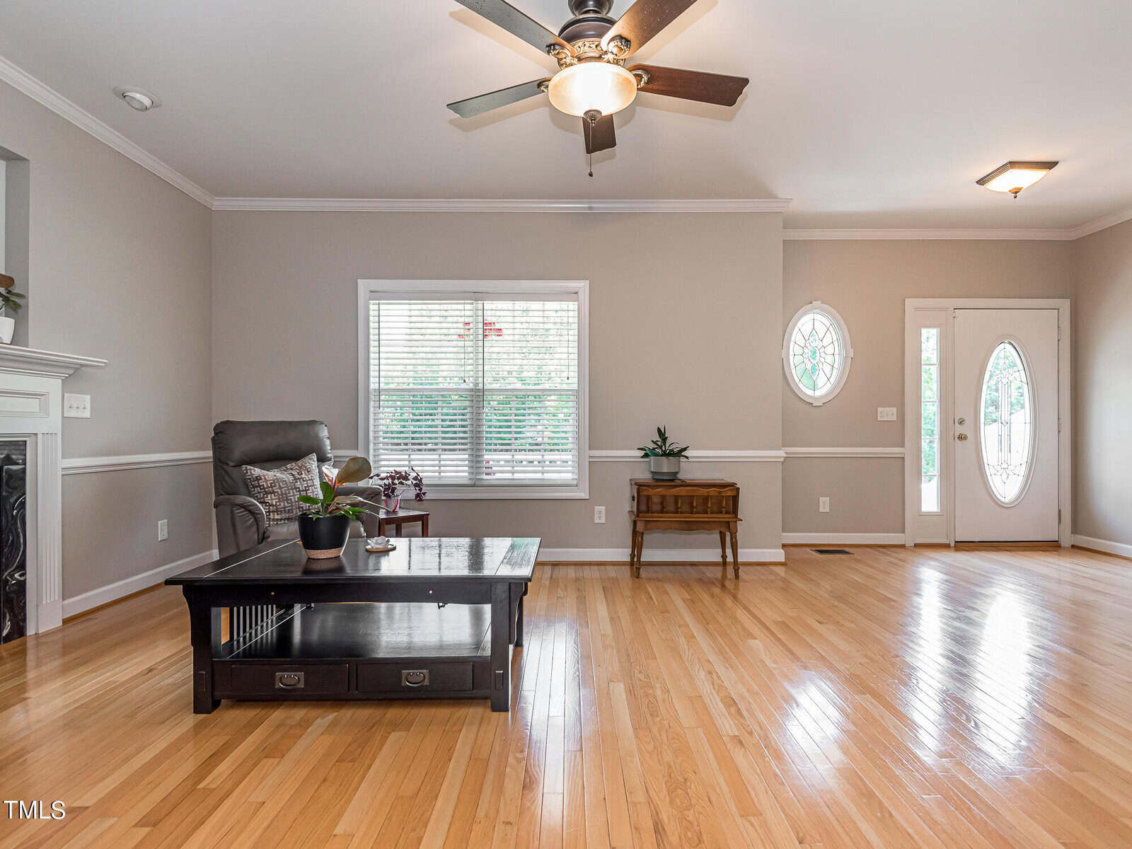 A spacious living room features light wooden flooring and neutral-colored walls. A ceiling fan with lights hangs above. The room includes a large window, a recliner, and a coffee table with plants. A decorative front door with an oval glass panel is visible.