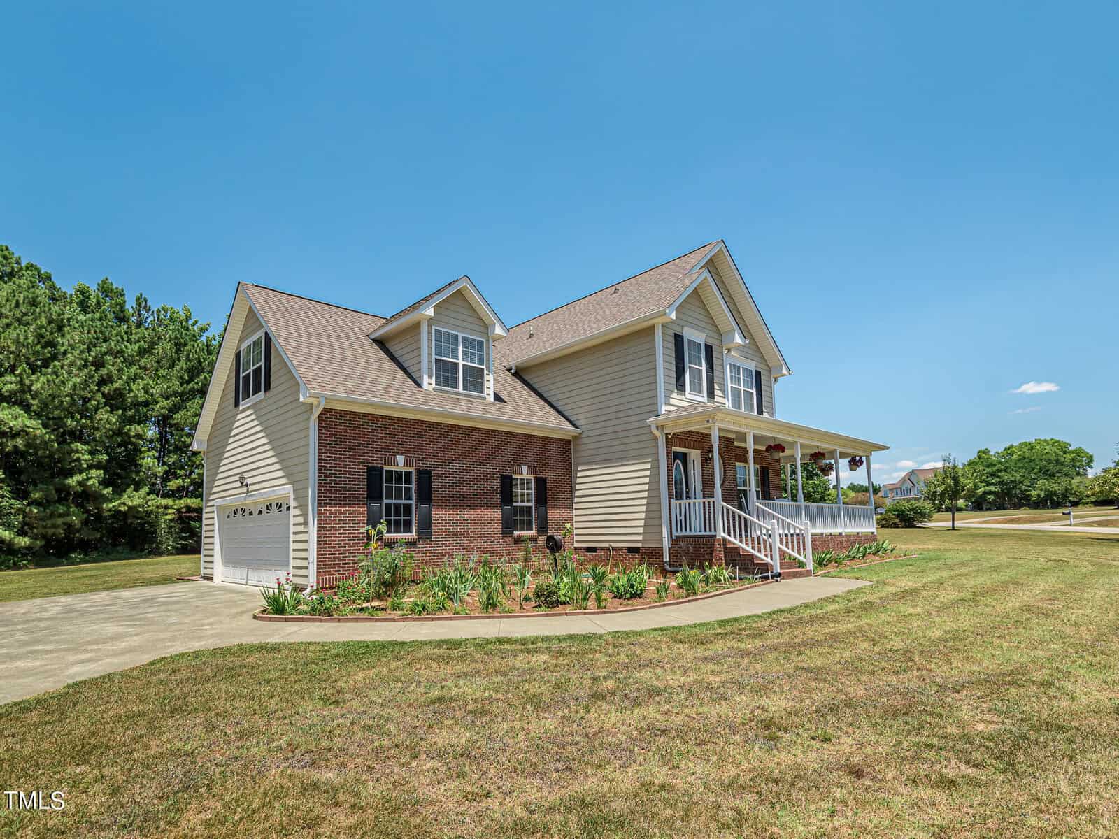 A two-story house with beige siding and red brick facade features a covered porch and attached garage. It is surrounded by a well-maintained lawn and bordered by trees under a clear blue sky.