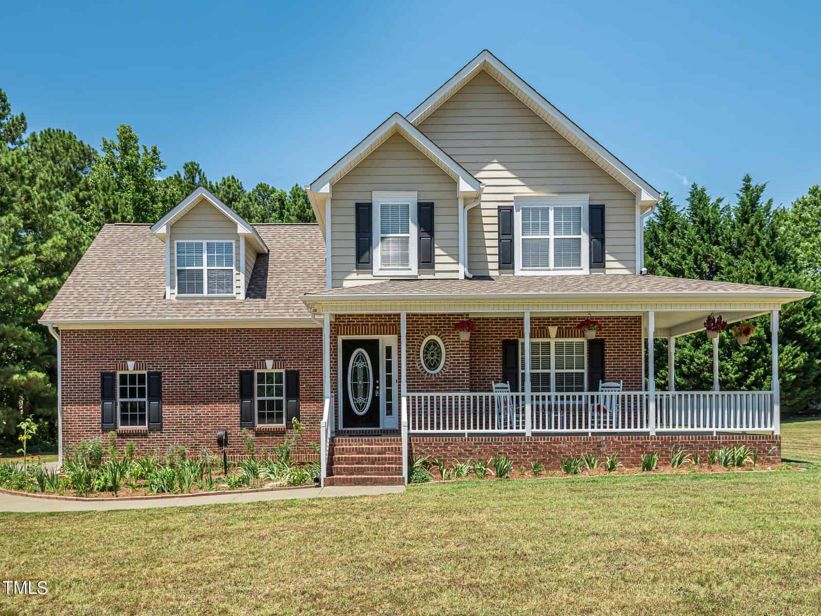 Located at 3080 Wren Road in Creedmoor, this two-story house boasts red brick and beige siding with a charming covered front porch featuring white railings. The home, adorned with multiple windows and black shutters, has a gabled roof and is surrounded by a manicured lawn with trees in the background.