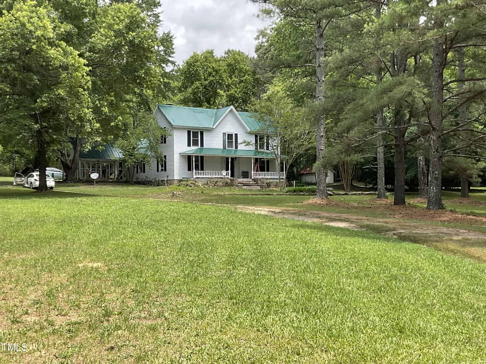 A large white two-story house with a green metal roof is nestled among tall trees with lush greenery surrounding it. A stone foundation supports the house, and a driveway leads up to the property. The scene appears serene and wooded, typical of a rural setting.