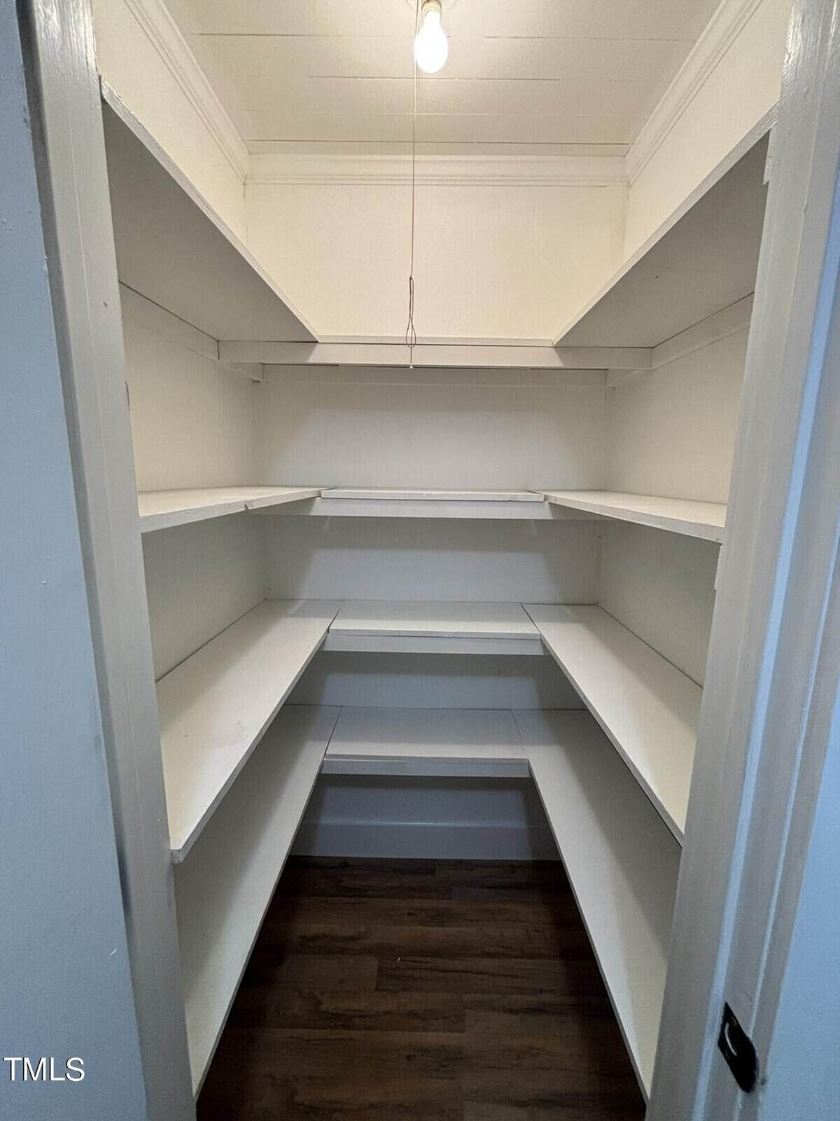 A pantry with white shelves on three walls, featuring multiple tiers for storage. The space is lit by a single exposed light bulb hanging from the ceiling. The floor is dark wood, and the walls are white. The door frame is seen in the foreground.