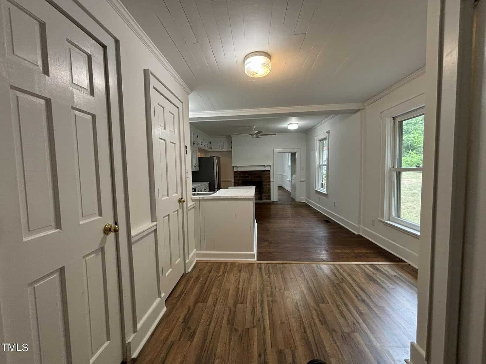 A view of a hallway leading to a kitchen and living area. The hallway has wood flooring and white paneled walls with two closed doors on the left. The kitchen area has white countertops and an island. Large windows in the living area provide ample natural light.