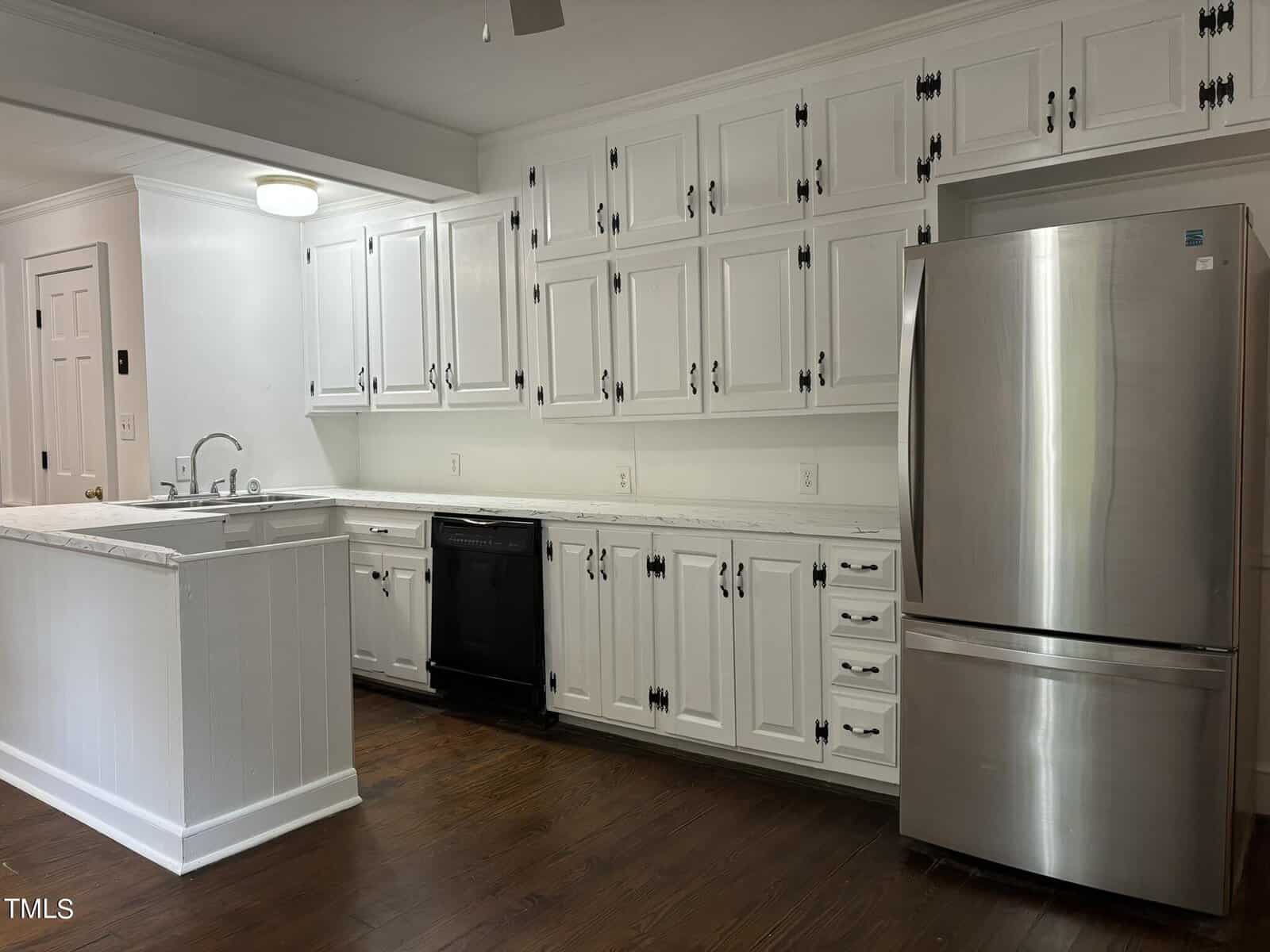 A modern kitchen featuring white cabinets with black hardware, a stainless steel refrigerator, and a built-in black dishwasher. The kitchen has white marble countertops, a deep sink, and wooden flooring. White walls and a ceiling light fixture complete the space.
