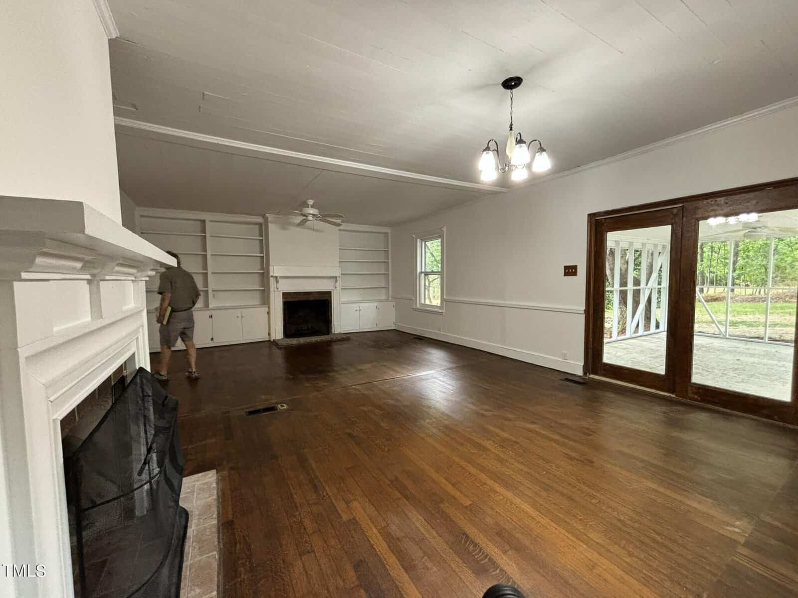 A spacious living room with hardwood floors and a white ceiling. The room features a fireplace with built-in shelves on either side, a ceiling fan, and a chandelier. Large glass doors lead to an outdoor patio. One person stands near the left side of the room.