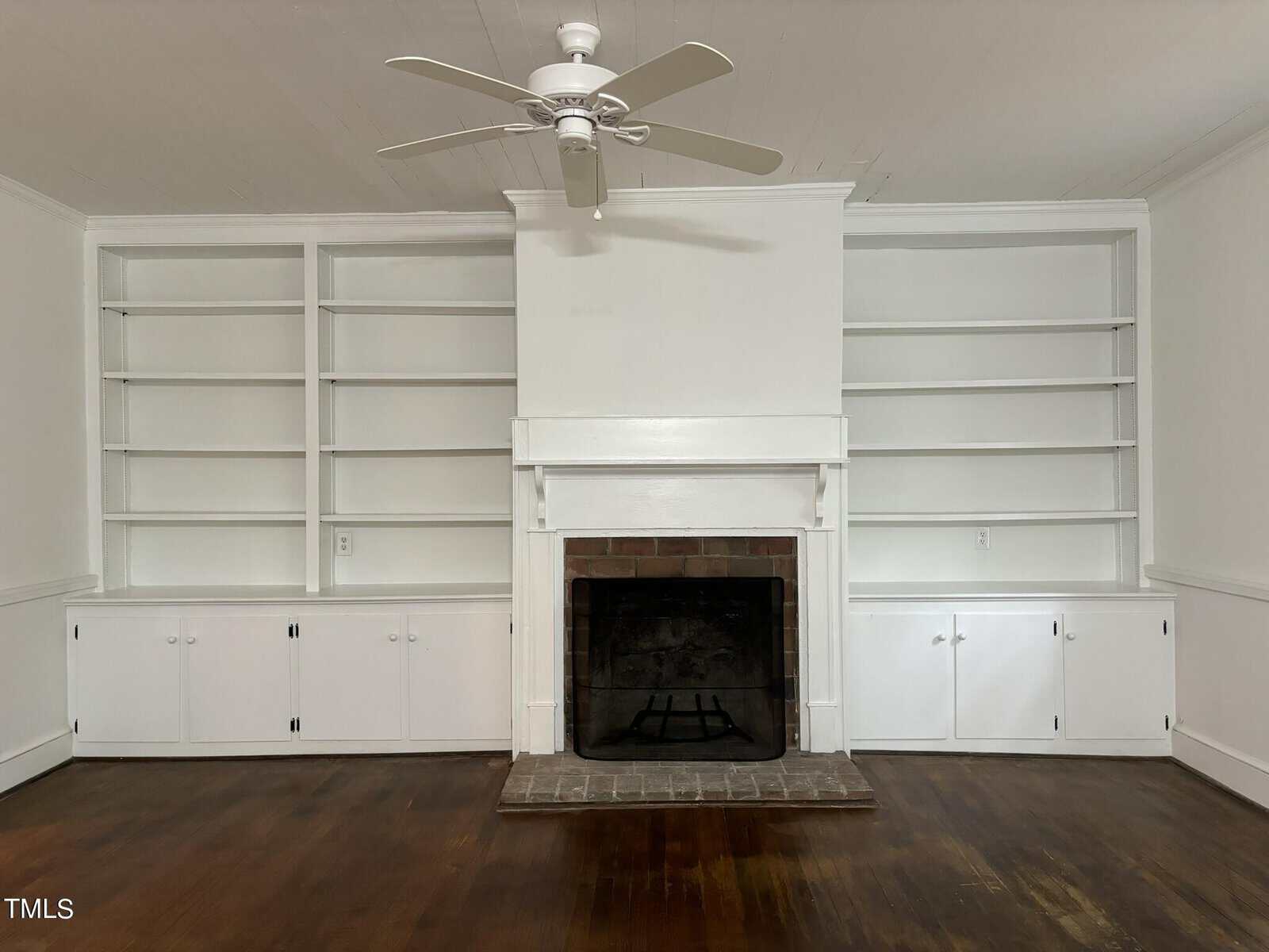 A well-lit, unfurnished living room with white walls featuring an empty fireplace at its center. Flanking the fireplace are built-in bookshelves and cabinets on both sides. A ceiling fan is mounted above the space. The wood floor is polished and in dark tones.