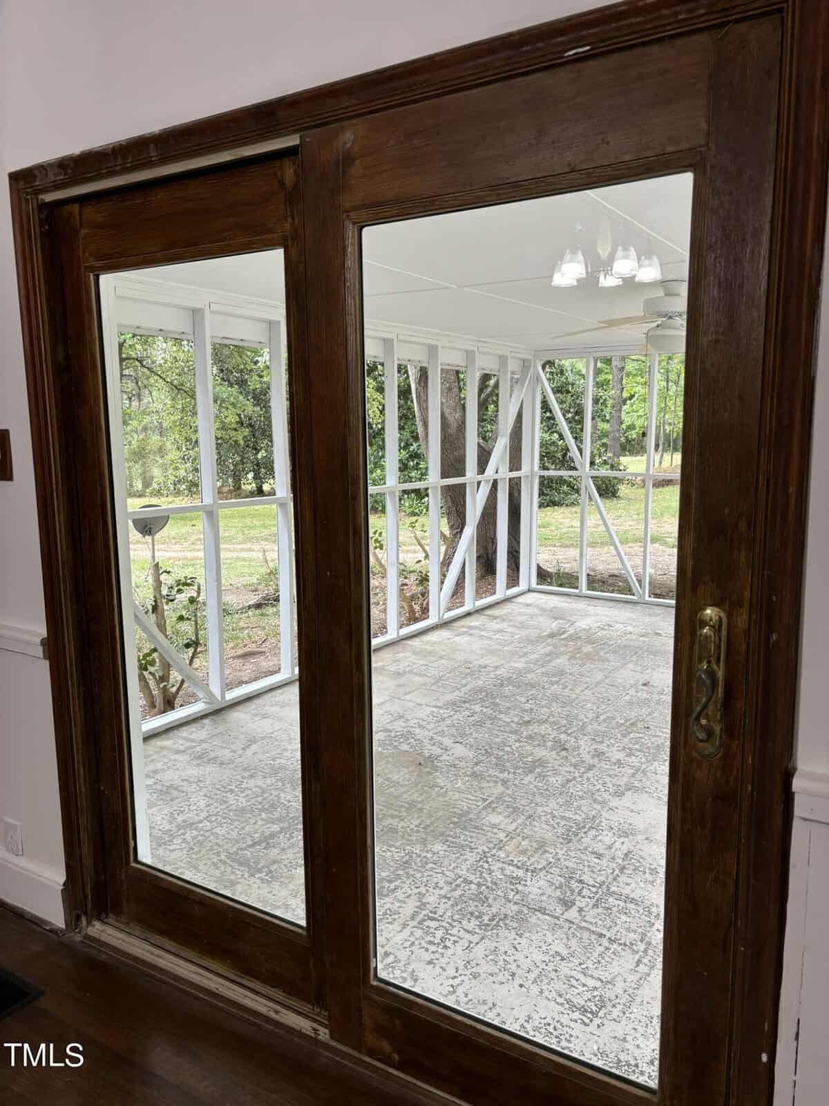 A wooden-framed sliding glass door opens to a screened porch. The porch has a white-framed structure with large windows providing a view of trees and greenery outside. The porch floor appears weathered and there is a ceiling fan with lights above.