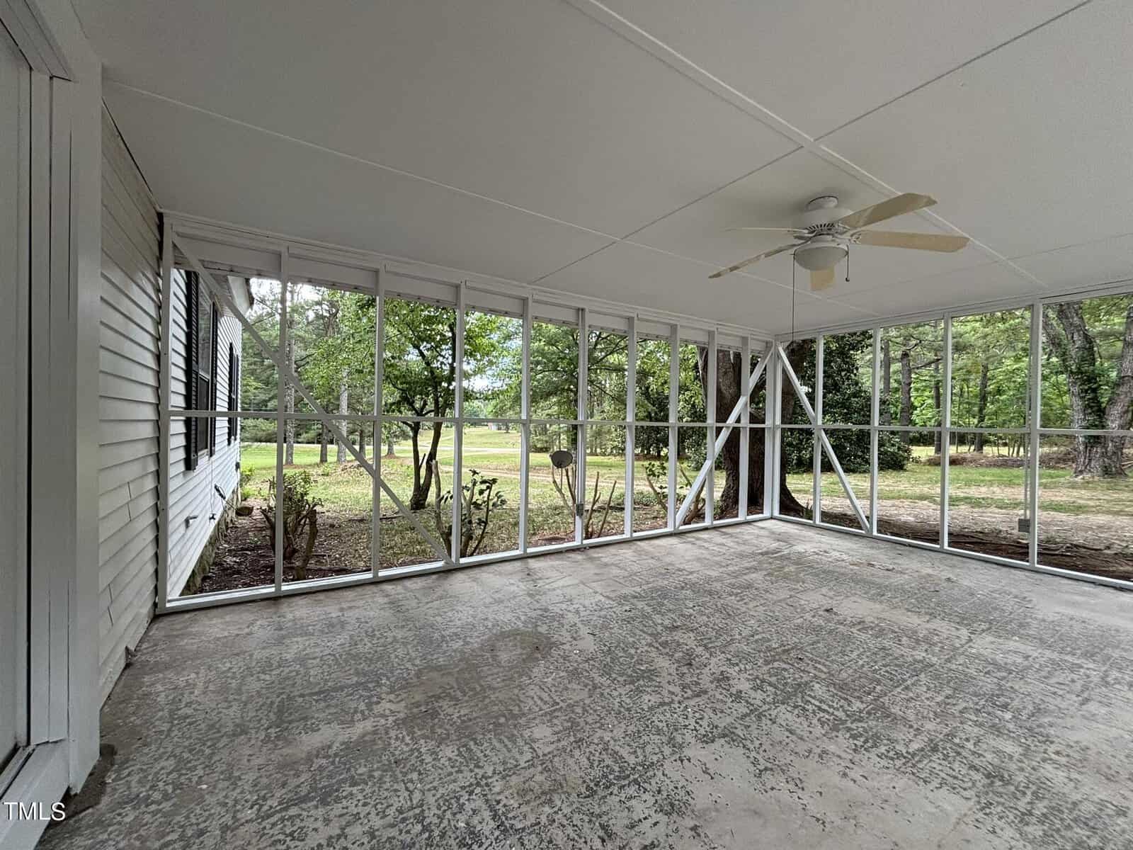 A spacious screened-in porch with a ceiling fan. The porch has metal framing and large screens providing a clear view of the surrounding green yard and trees. The floor is made of concrete, and the space appears clean and well-maintained.