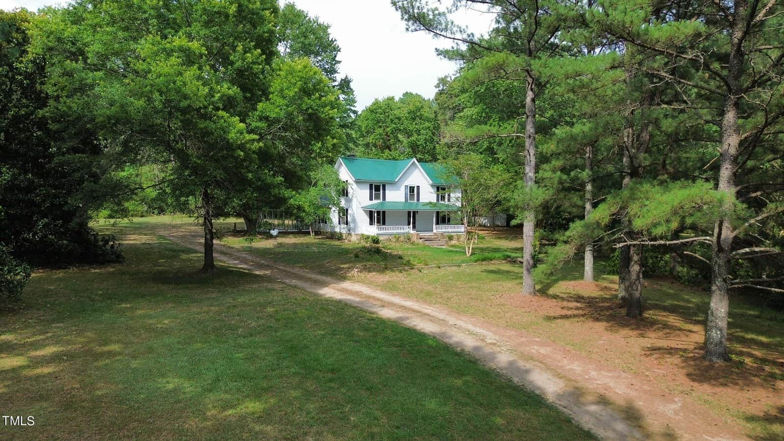 A white two-story house with a green roof at 1734 Rocky Ford Road is surrounded by numerous trees. There is a dirt path leading up to the house, which has a front porch. The area in Kittrell is lush with greenery and appears serene and secluded.