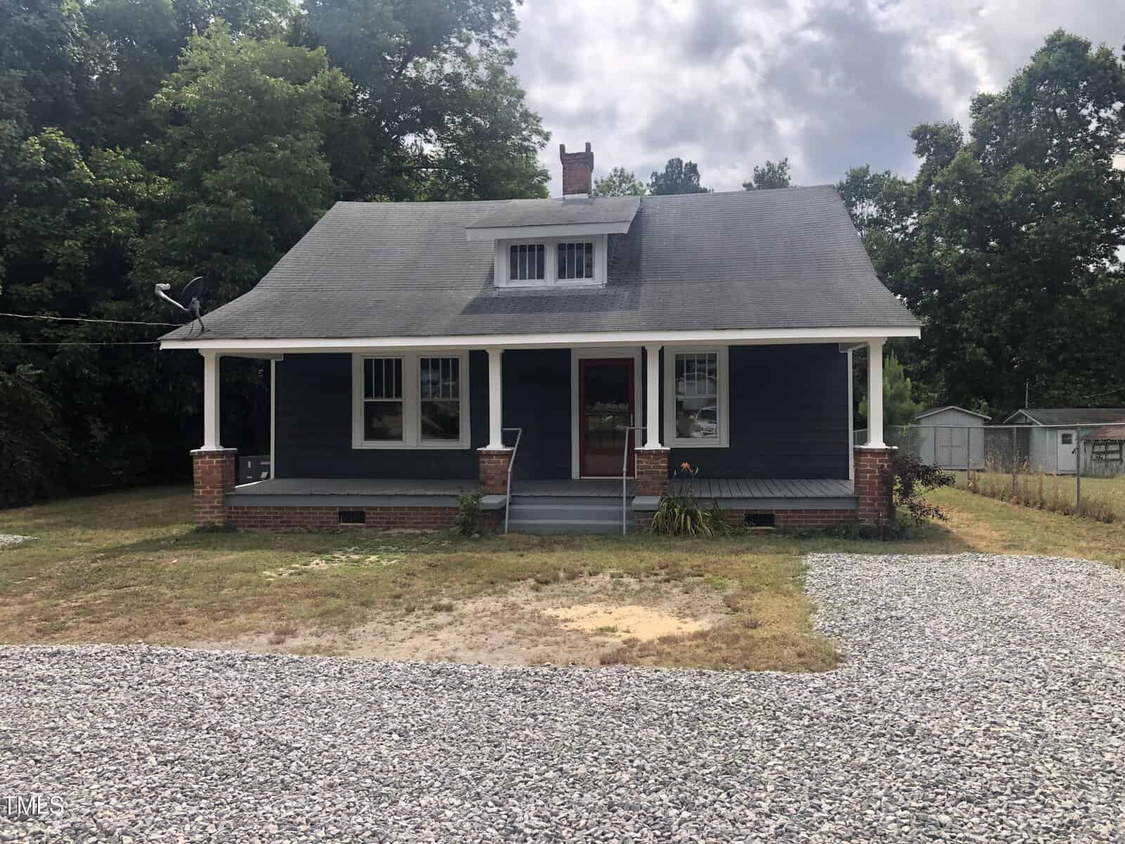 A charming single-story house located at 3124 Raleigh Road, Henderson NC, featuring dark blue wooden siding, a steeply pitched roof, and a small upper window. The front porch is supported by white columns and brick steps. Surrounded by a gravel driveway and a grassy yard with trees in the background.