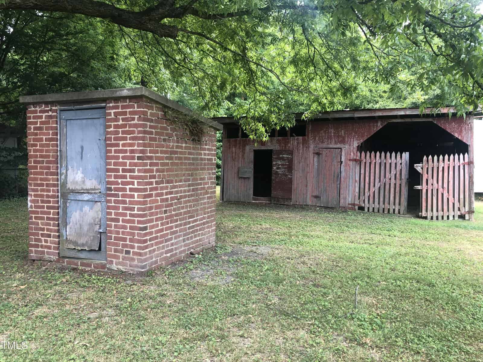 A weathered red barn with a gabled roof and open stalls stands partially surrounded by a white picket fence. In the foreground, there is a small, old brick shed with a wooden door. Lush green grass and leafy trees complete the scene.