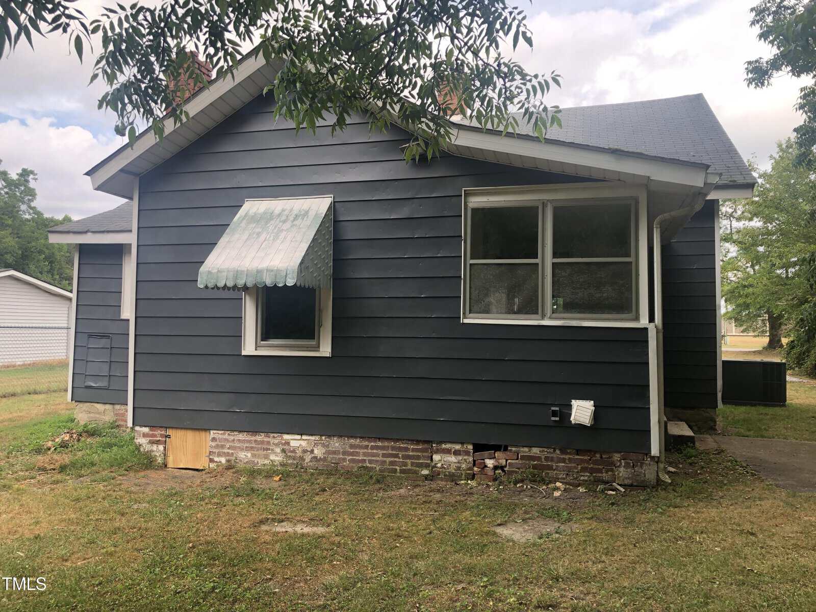 A small blue house with a sloped roof, a covered front window, and visible white window frames. The base is brick, and the grass around the house is sparse. Trees frame part of the image, and the sky is cloudy. The house appears older with some wear and tear.