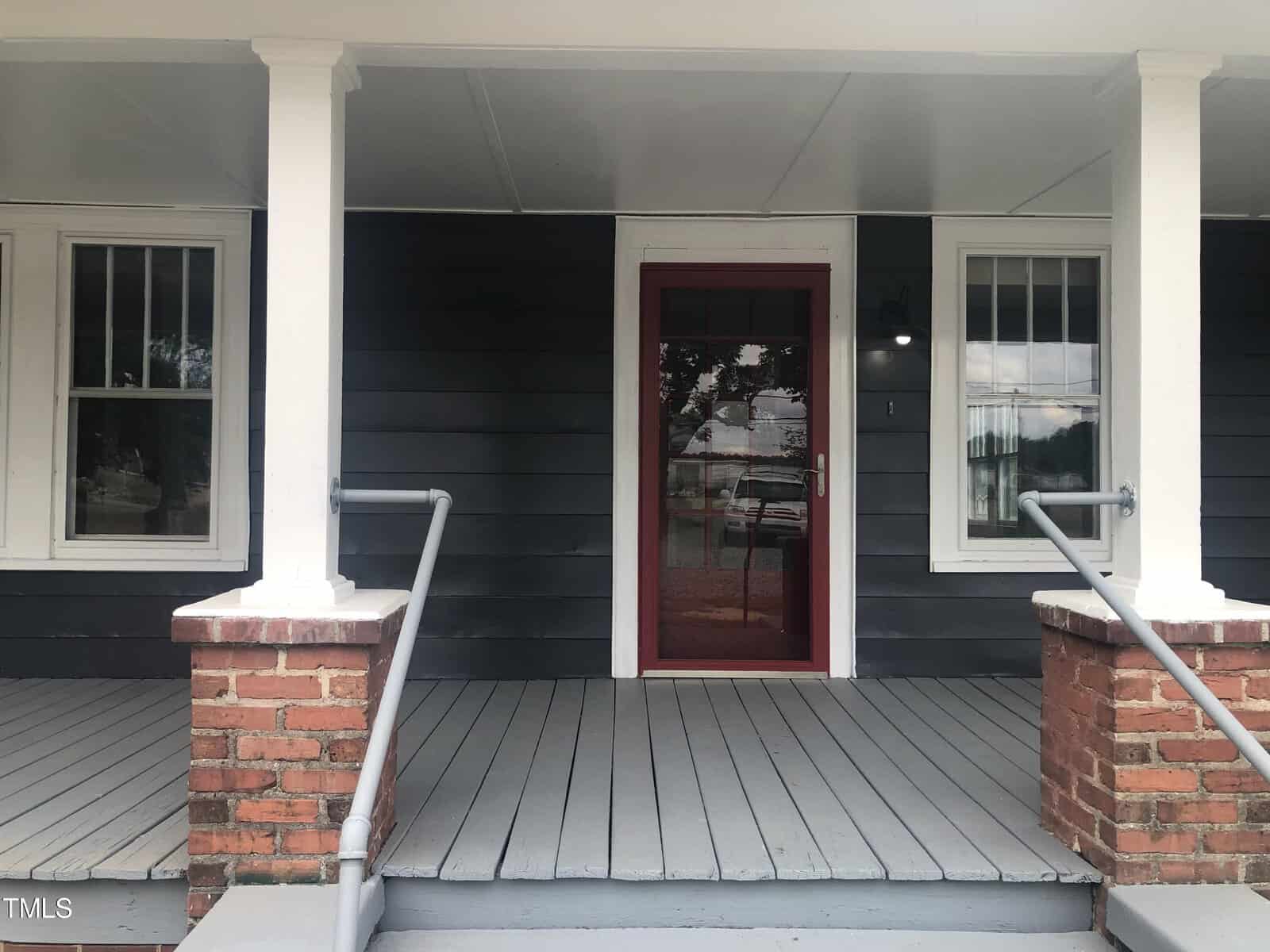 A house entrance with a red door and two flanking white-framed windows. The porch has gray flooring, a white ceiling, and white pillars resting on brick bases. The steps to the porch are also gray and have metal railings on both sides.