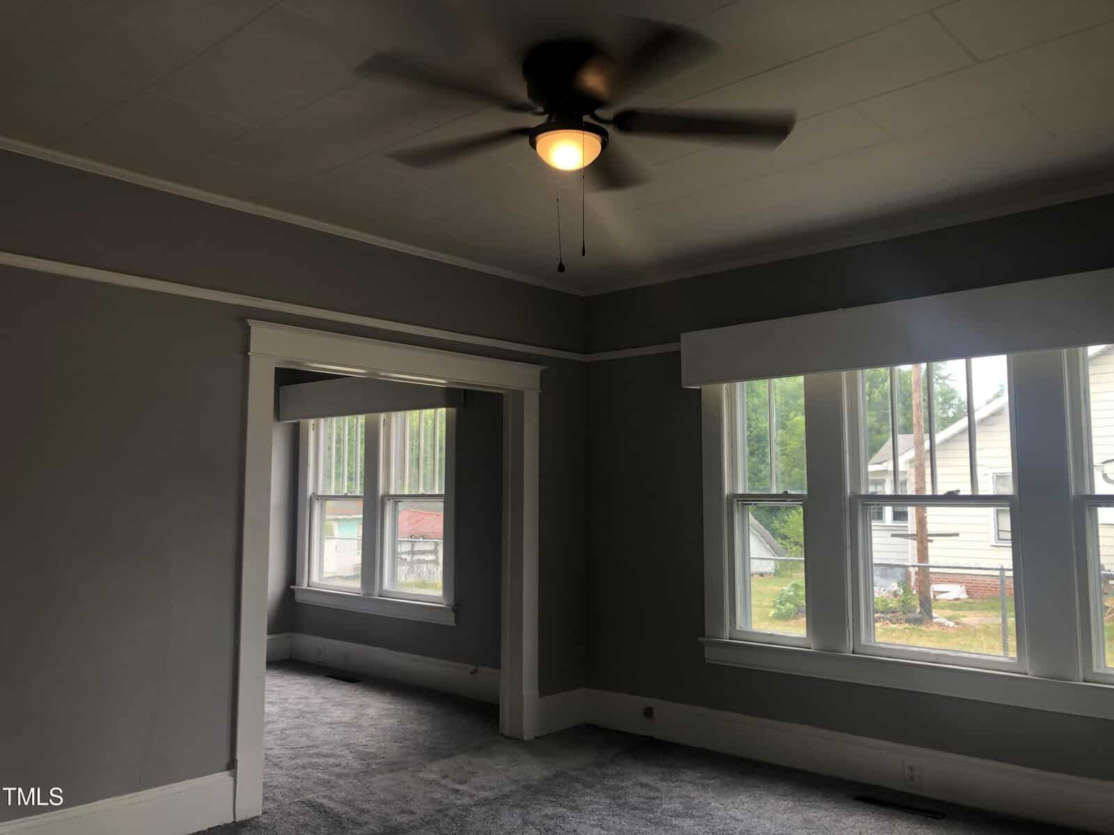 A modern, unfurnished room with gray walls, white trim, and large windows letting in natural light. The room features a ceiling fan with a light and gray carpet. An open doorway leads to another similar room with more windows.