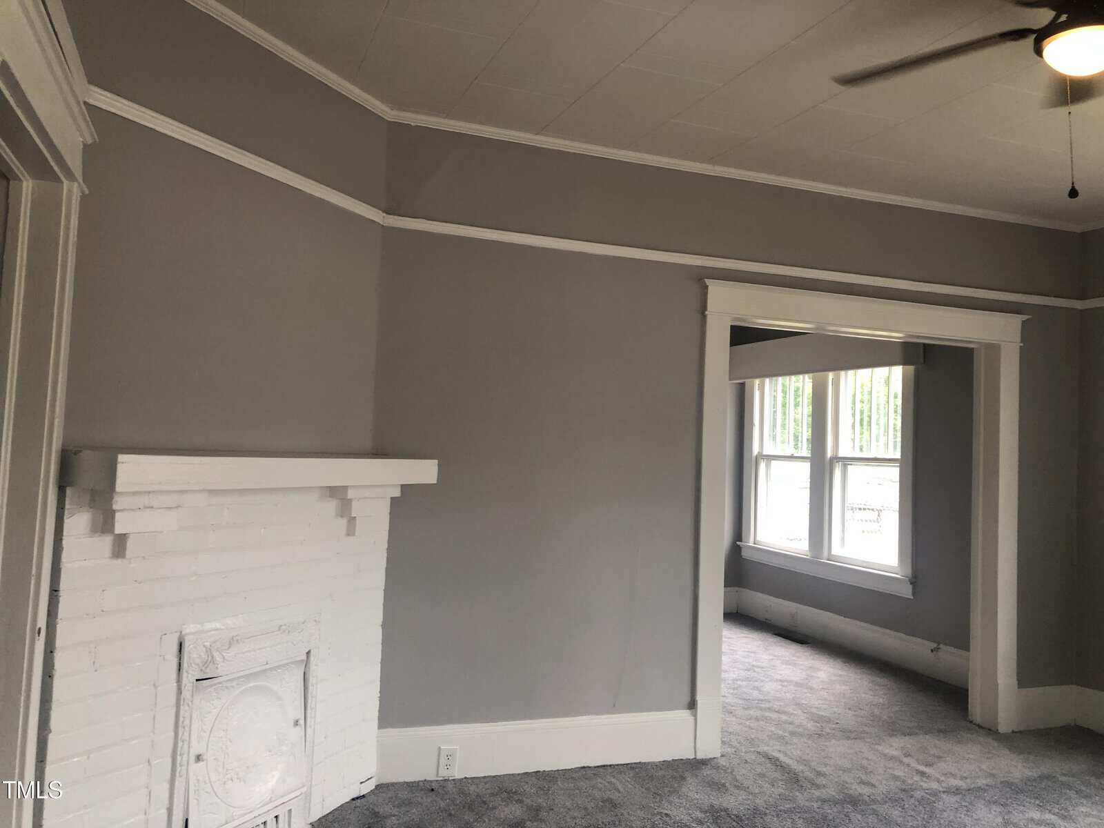 A gray-walled room with white trim featuring a white-painted brick fireplace. The room has a ceiling fan, and an adjoining space with large windows letting in natural light. The floor is carpeted in a light shade of gray.