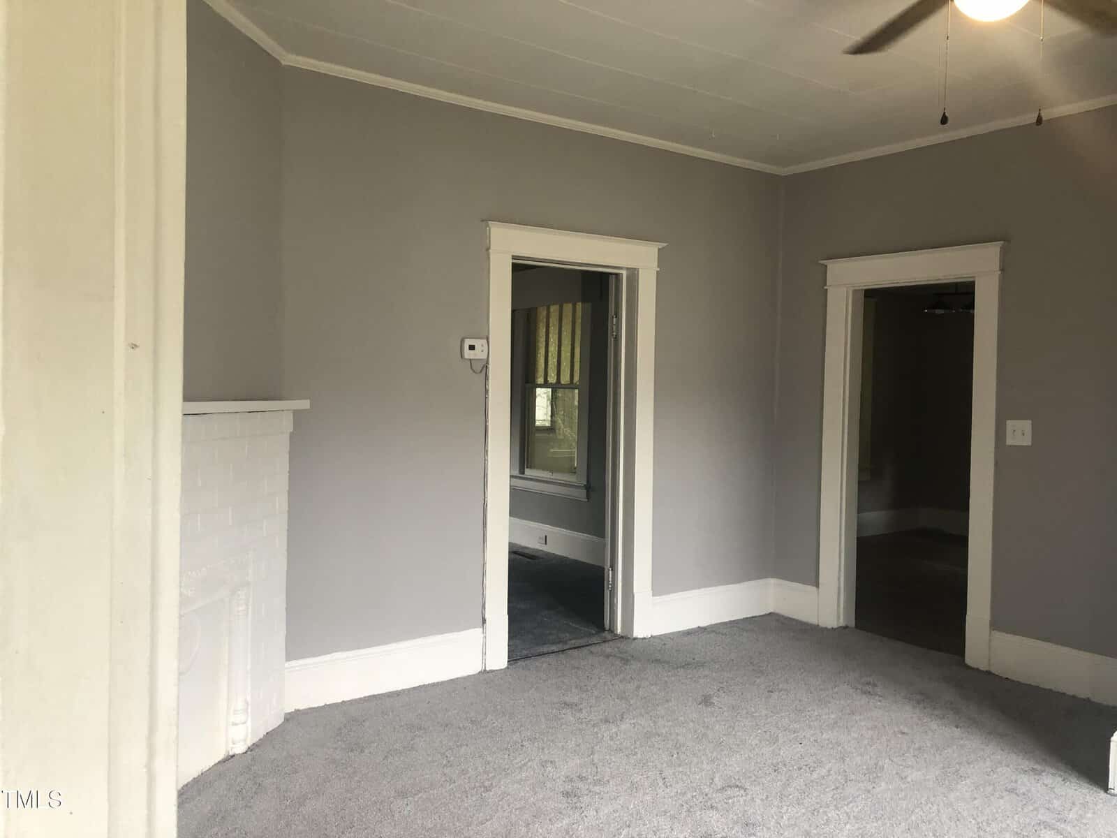 A room with grey walls and carpeting, featuring white trim, a white painted brick fireplace on the left, and two doorways: one leading to another room and one to a hallway. A ceiling fan with light is partially visible in the top right corner.