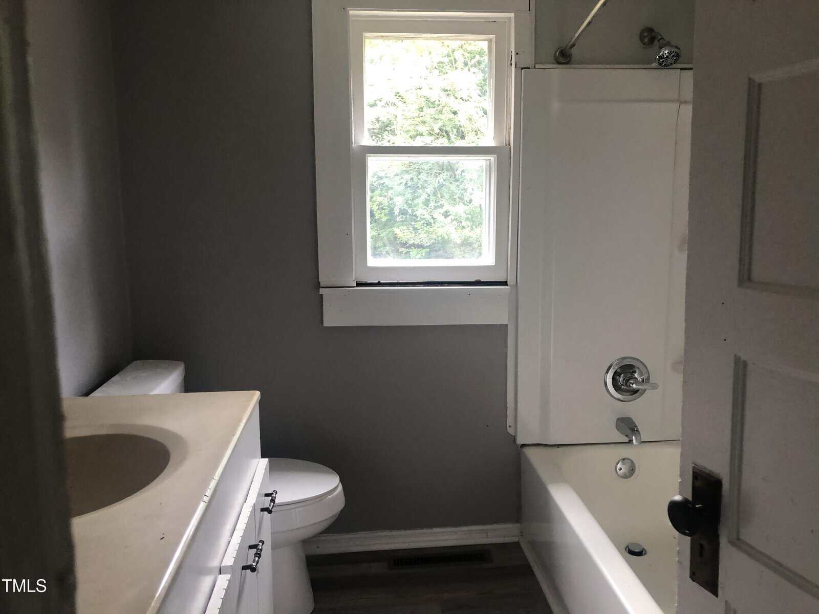 A small bathroom with gray walls features a white bathtub-shower combo, a white toilet, and a white sink with a light-colored countertop. Above the bathtub is a window with a view of greenery outside. A door is partially open, revealing the room.