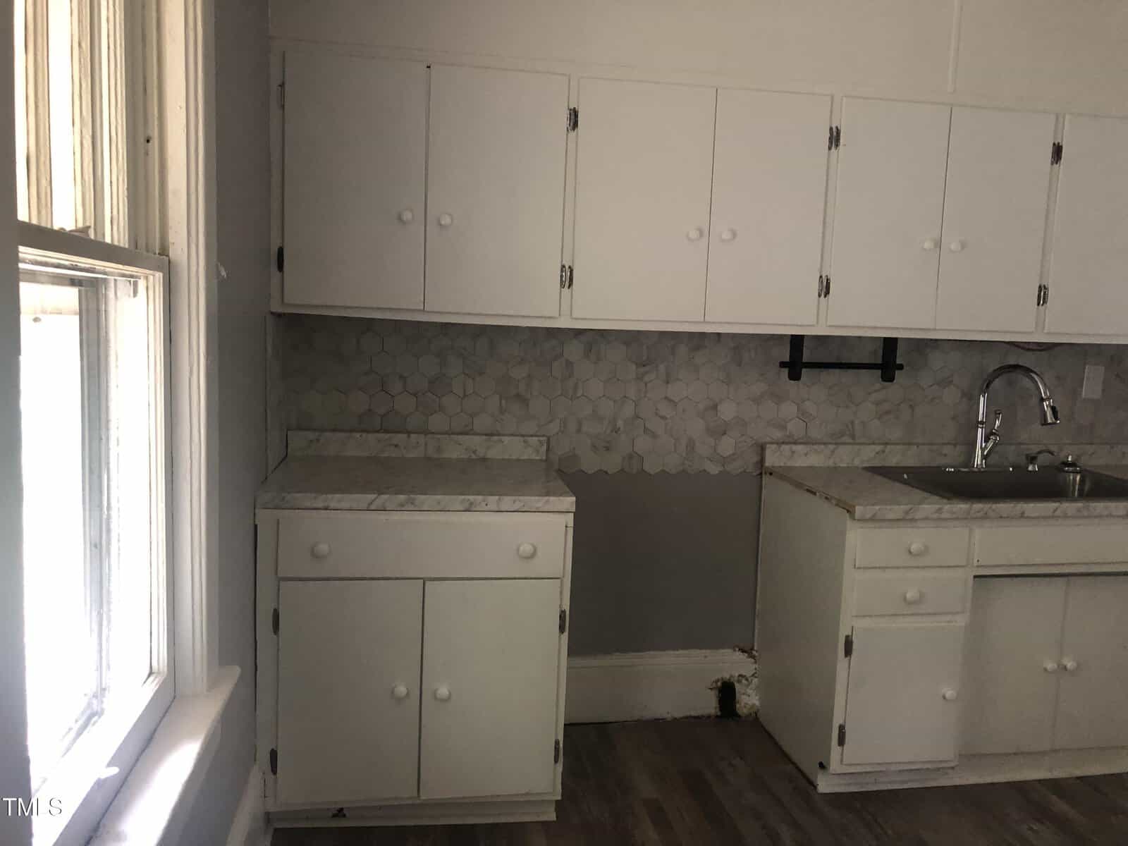 A kitchen with white cabinets and countertops. The backsplash area is partially incomplete, showing exposed wall. There is a sink with a faucet on the right side of the image, and a large window on the left, allowing natural light into the room.