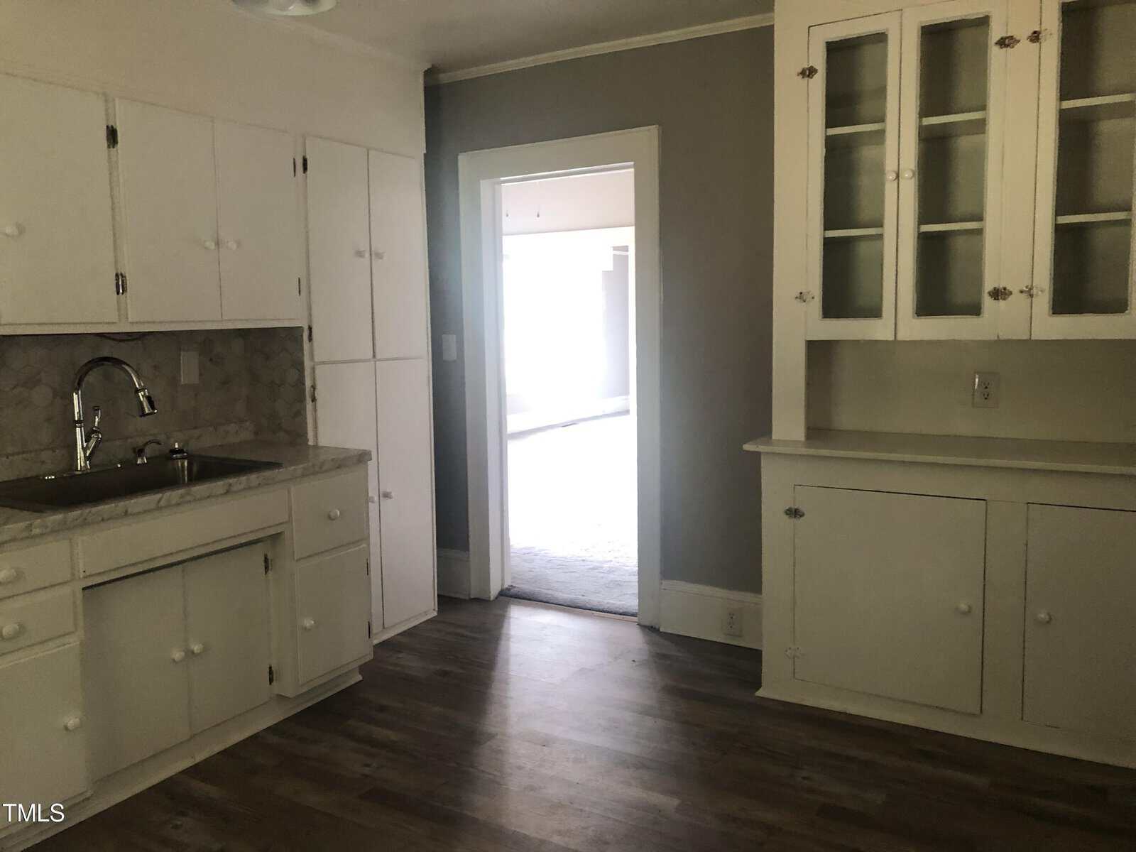A vintage kitchen features white cabinetry with glass-paneled upper cabinets, light gray walls, and wooden flooring. There is a single-basin sink with a backsplash on the left. The room leads to another sunlit room through an open doorway.
