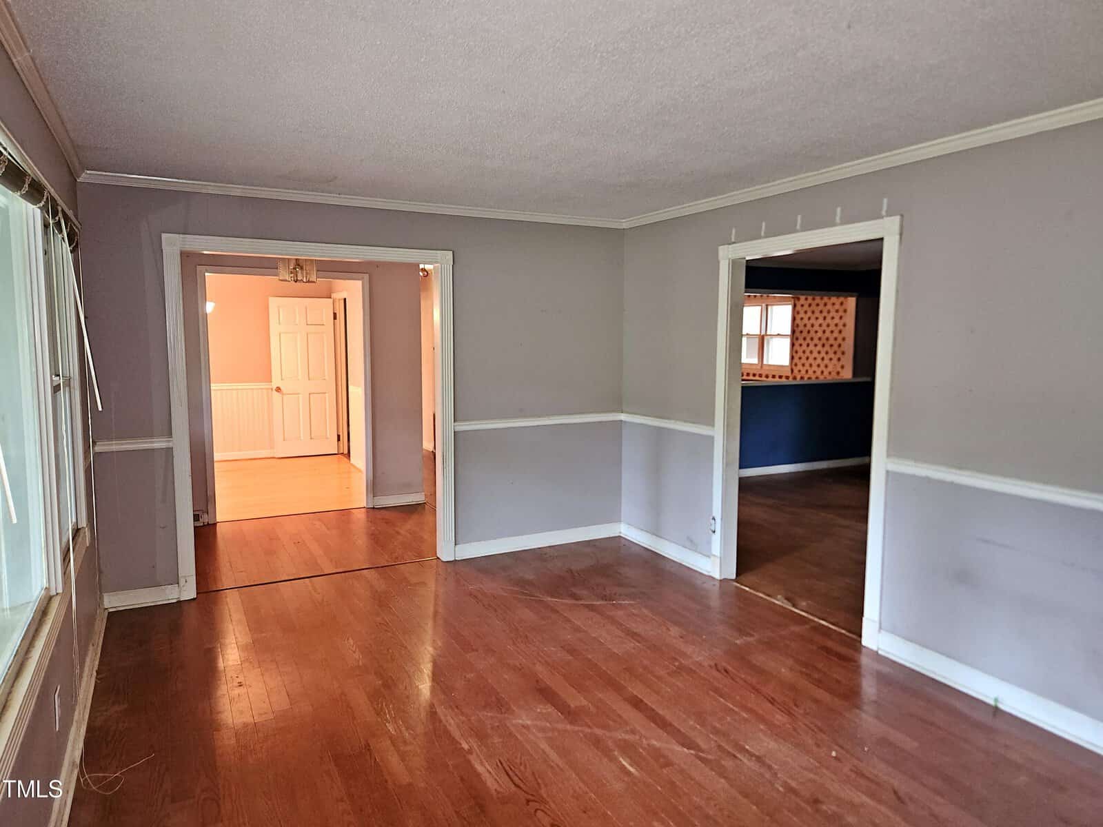 An empty room with light gray walls and wooden floors. A large window is on the left, with adjoining doorways leading to other rooms. One room is brightly lit and another is darker.