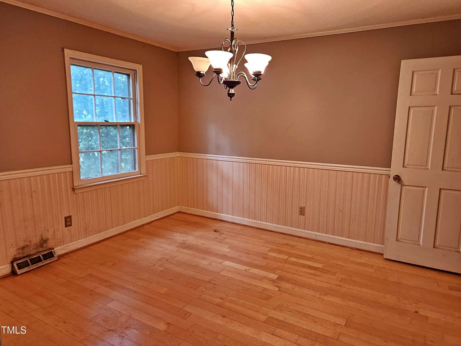 A small, empty room with light wood flooring and beige walls featuring a chair rail. A window with a view of greenery is on the left wall. A white door is partially open on the right, and a ceiling light fixture hangs in the center.