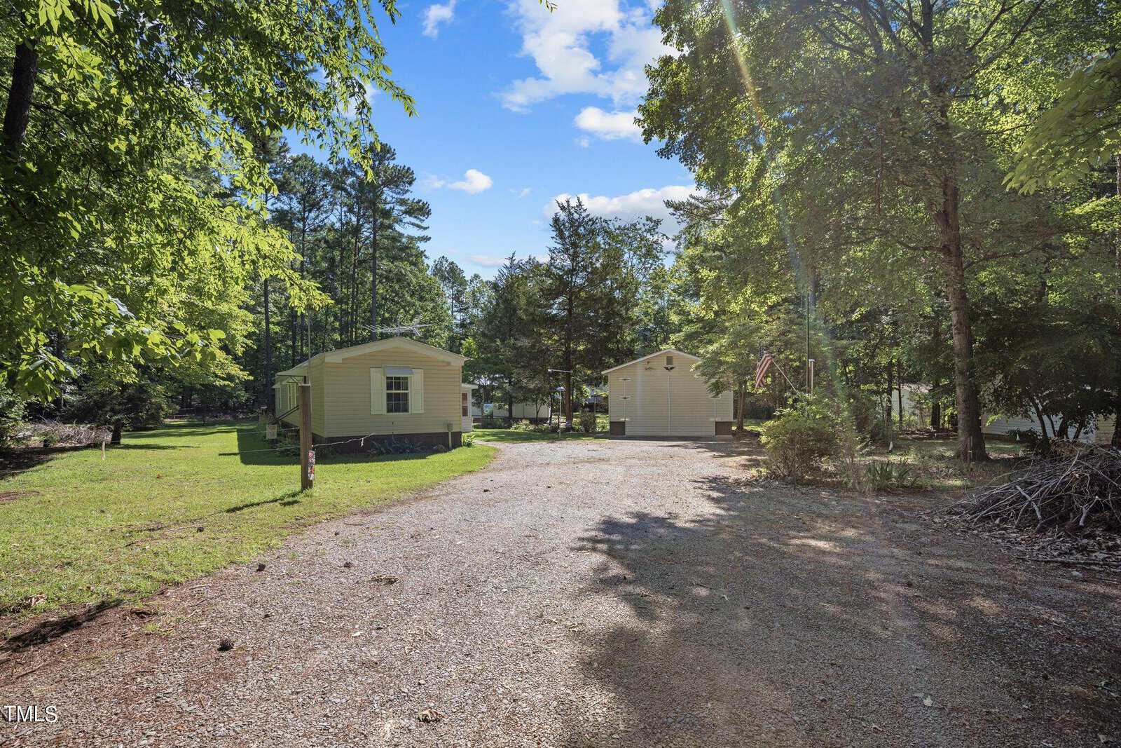 A gravel driveway leads to two light-colored mobile homes, surrounded by green trees and grass. The driveway is dappled in sunlight, and an American flag is visible in front of one of the homes. The sky is blue with scattered clouds.