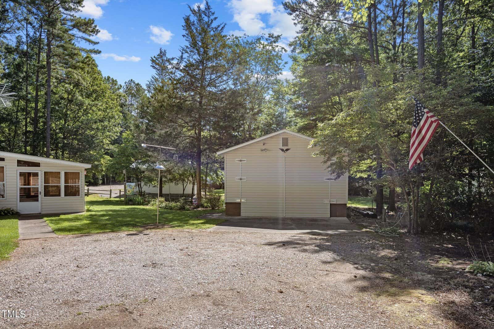 A gravel driveway leads to a small yard with a white storage building and a house on the left. The yard is surrounded by tall trees and includes a flagpole with the American flag. The sky is clear with some clouds.