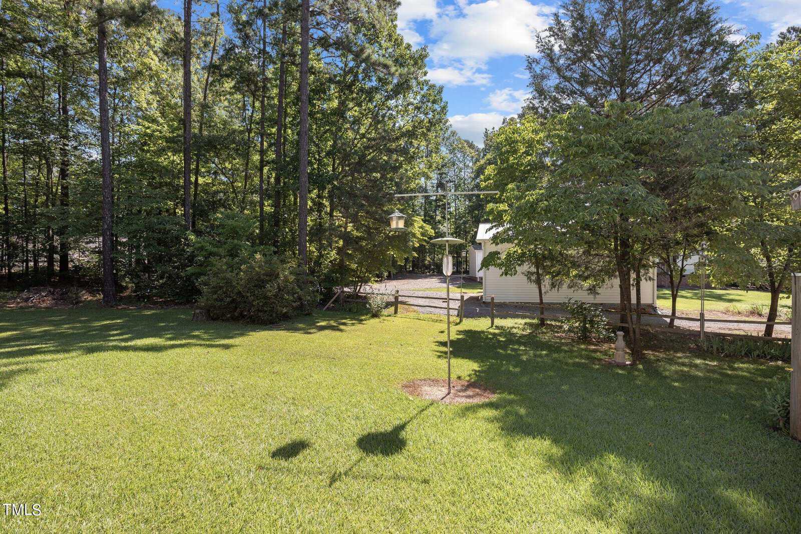 A well-manicured grassy backyard surrounded by tall trees. A birdhouse stands in the center, casting a shadow on the lawn. In the background, there is a light-colored shed with a small porch, and a gravel path leading to it. The sky is partly cloudy.