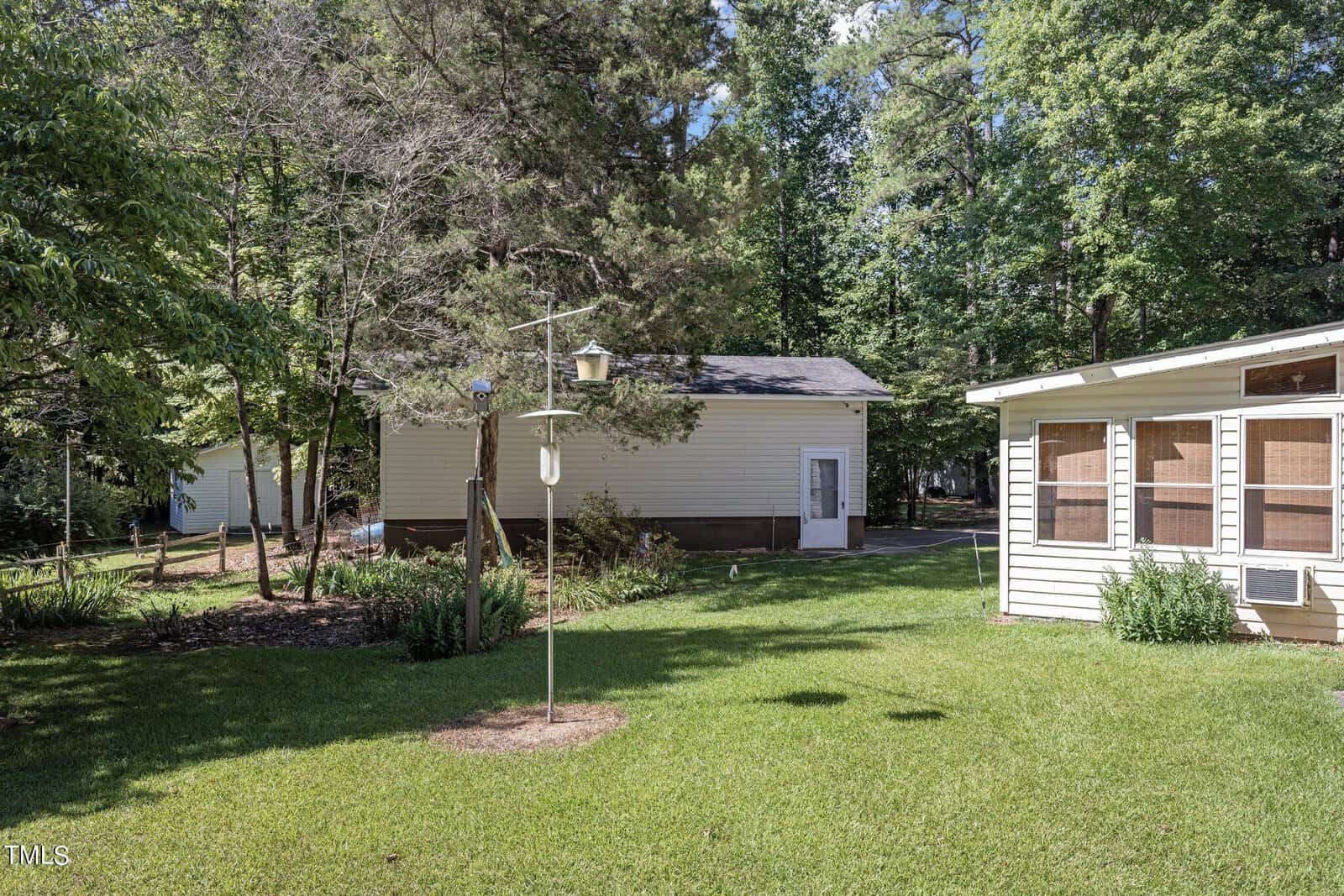 A backyard with a well-maintained lawn, a few trees, and a bird feeder hanging from a pole. There is a small beige building surrounded by trees in the background, and a larger building with screened windows on the right. The scene is bathed in sunlight.
