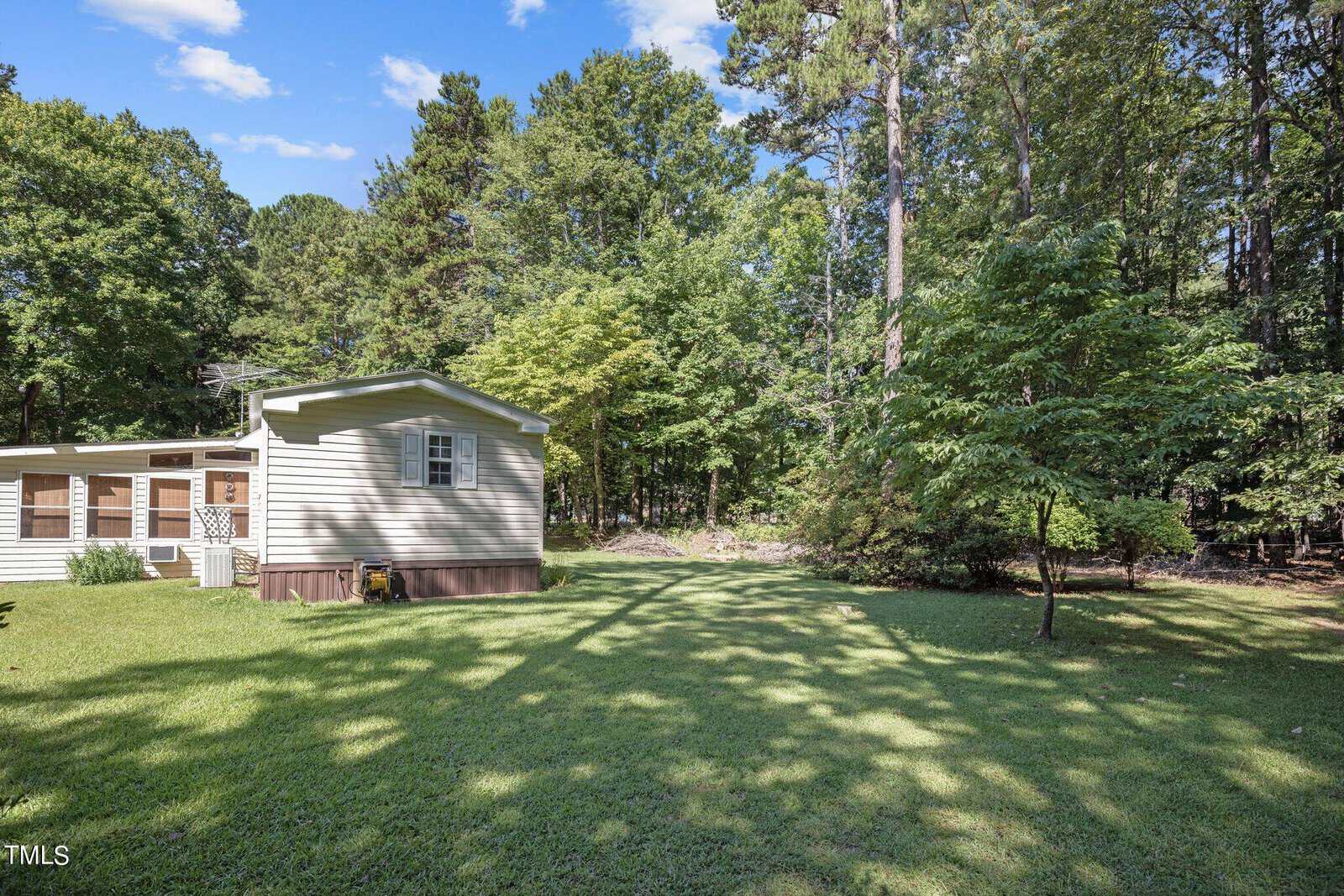 A beige mobile home stands in a green yard, surrounded by tall trees. The home features a small porch with windows, and the yard is well-maintained with lush grass extending to the edge of a dense forest. The sky is clear and blue with a few scattered clouds.