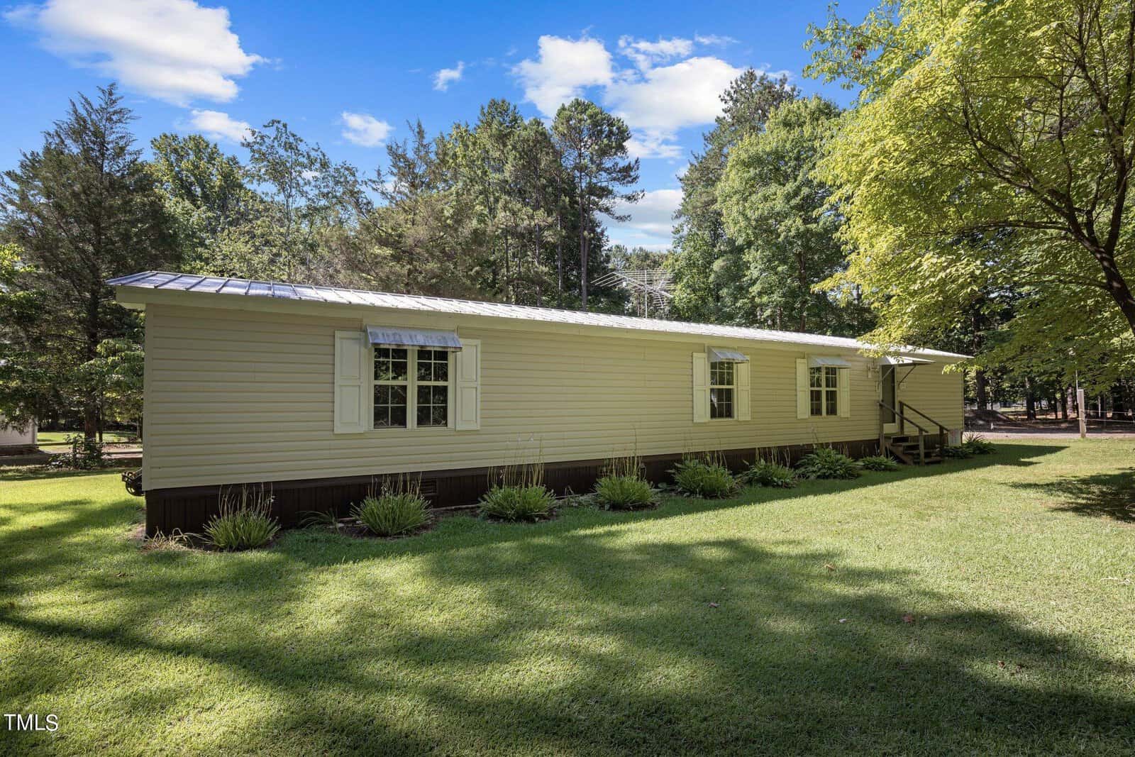 A beige-colored single-wide mobile home with white shutters and a brown skirt sits on a well-maintained grassy lawn at 15 Waters Edge Loop Rd in Henderson, NC. Tall, dense trees surround the property, and the sky above is partly cloudy on a sunny day. Steps lead to the front entrance on the right side.
