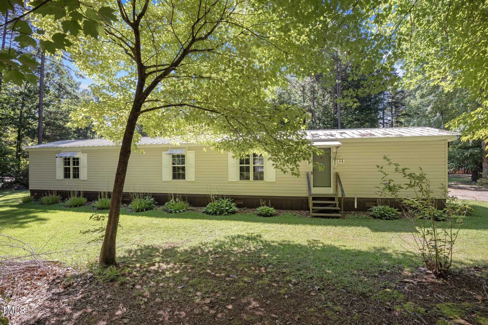 A single-story, beige mobile home with a metal roof is situated in a wooded area. The front yard has green grass, a few bushes, and a large tree providing shade near the front door, which is accessed by a small set of steps.